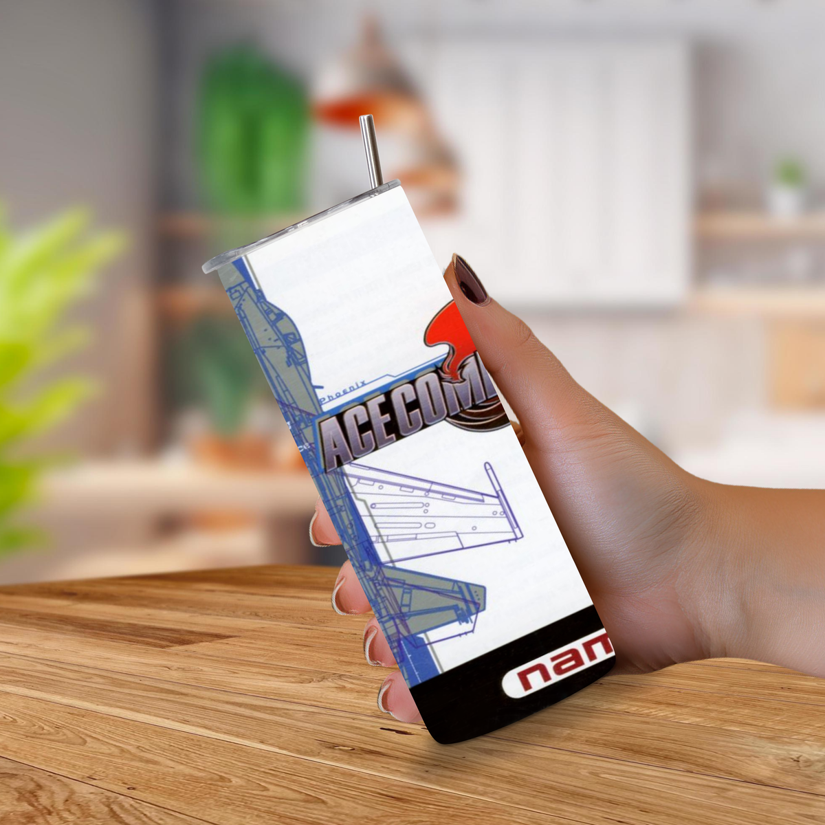 A hand holding a 20oz tumbler of PS1 Ace Combat 2 themed design and a metal straw, set against a blurred indoor background with a wooden table.