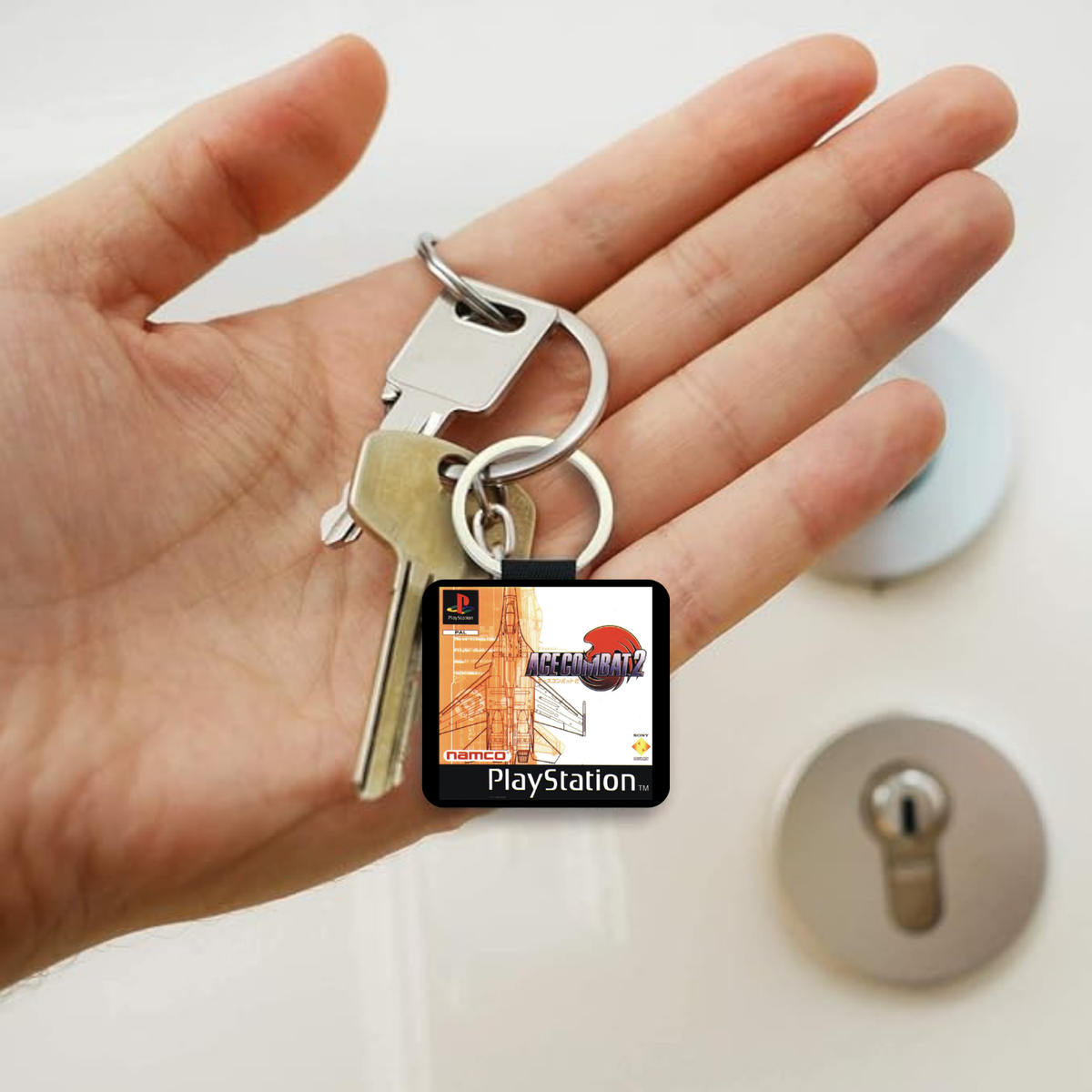 A hand holding a small square PlayStation Ace Combat 2 keychain with attached keys, shown in front of a lock cylinder.