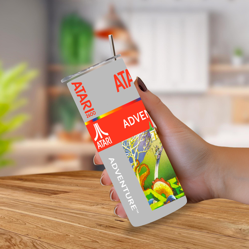 A hand holding a tall, slim Atari Adventure tumbler with a straw, set against a blurred kitchen background.