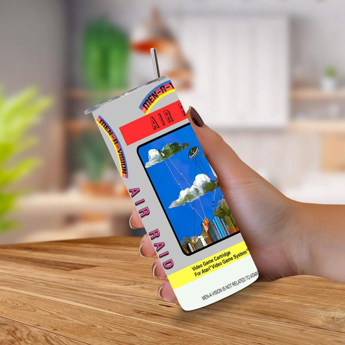 A hand holding a novelty 20oz tumbler designed to look like a vintage Atari air raid cartridge, featuring a colorful illustration of a blue sky with clouds and a city skyline, set against a blurred indoor background.