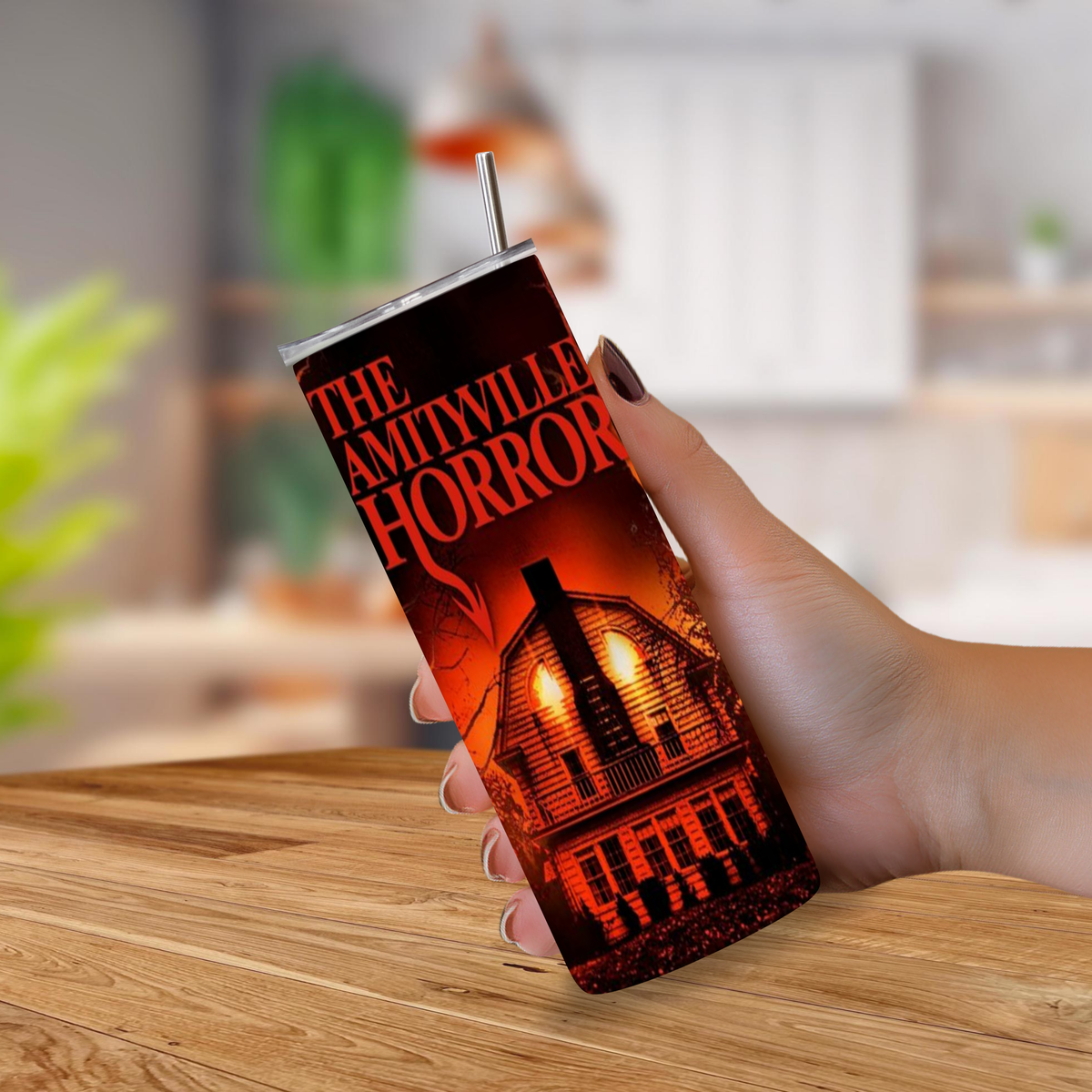 A hand holding a tall insulated tumbler with a straw, printed with the title 'The Amityville Horror' and a dark haunted-house illustration, set against a blurred indoor background.