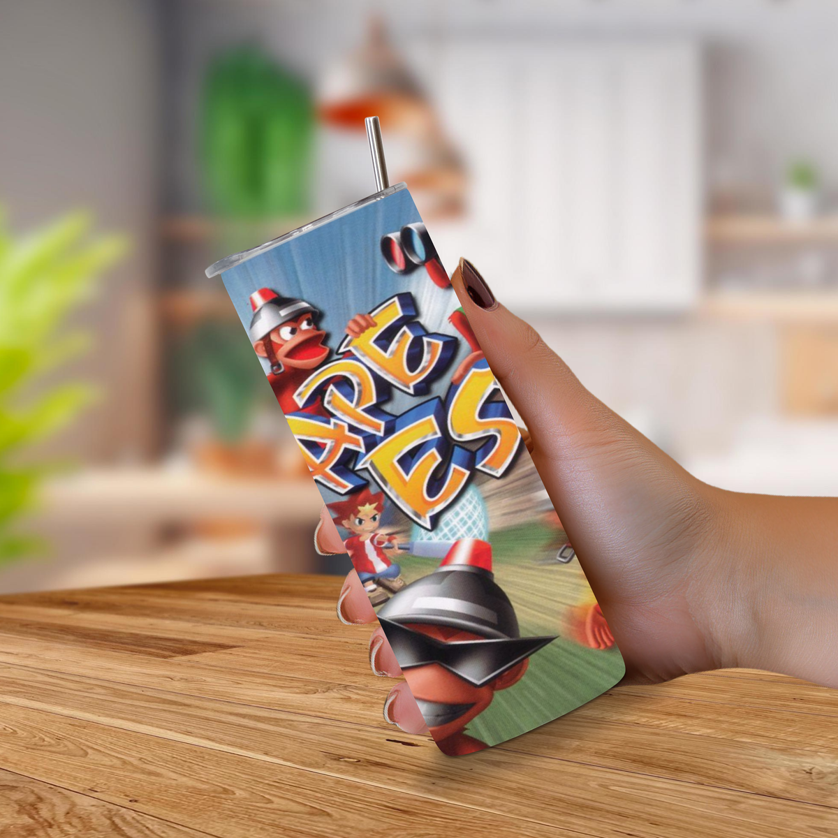 A hand holding a 20oz tumbler featuring a colorful cartoon graphic with the word 'Ape Escape' in large letters, set against a blurred indoor background with a wooden table in the foreground.