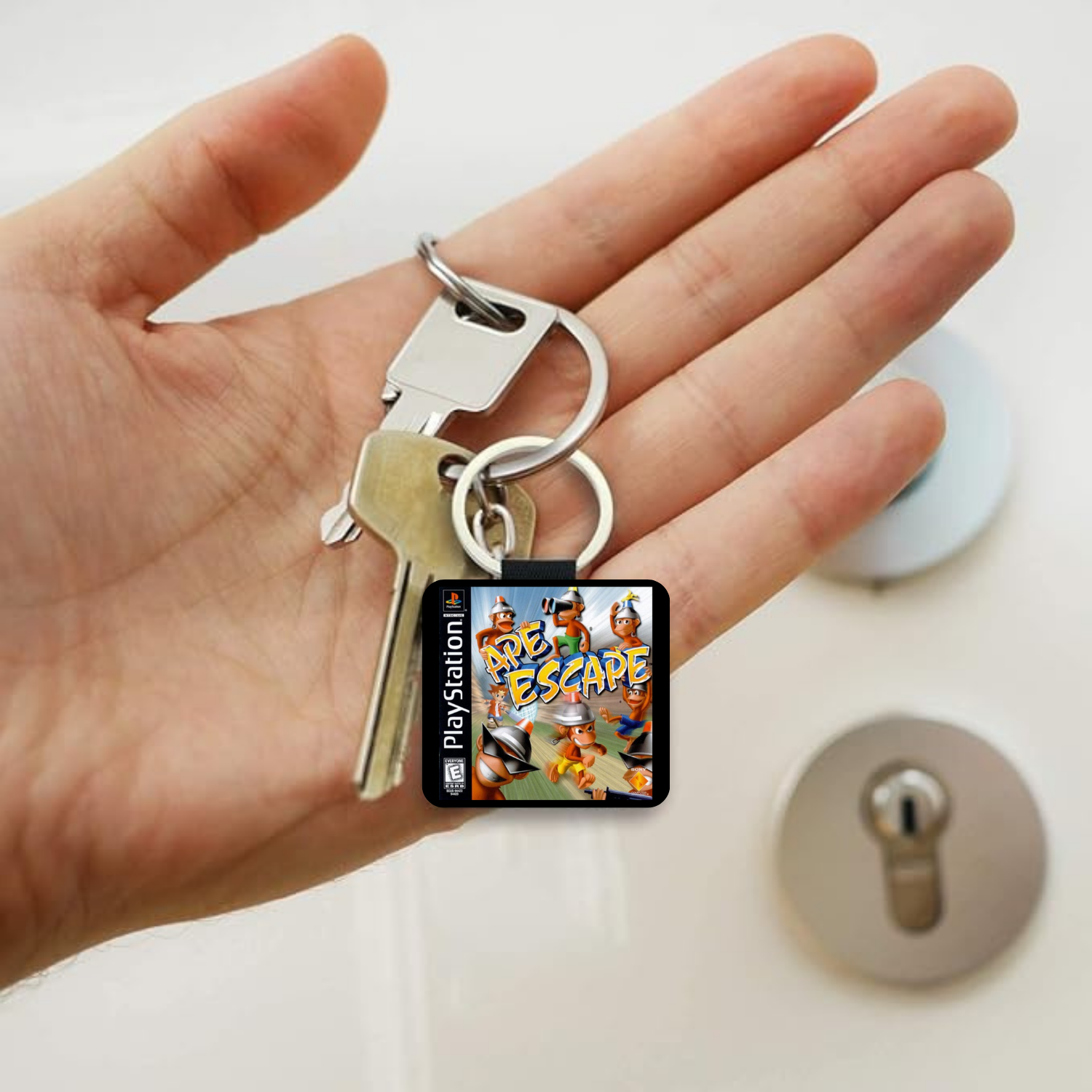 A hand holding a silver keyring with two keys and a small square PlayStation-themed charm featuring colorful cartoon graphics and the text 'Ape Escape'.