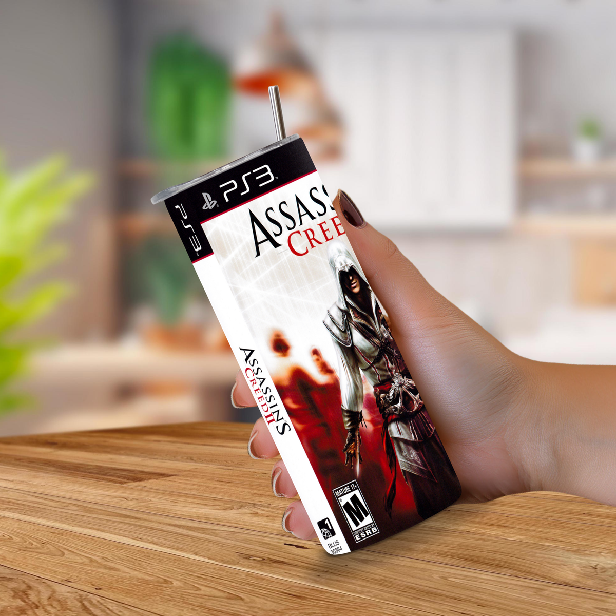A hand holding a PS3-shaped game cover designed to look like a 20oz stainless steel tumbler, featuring Assassin's Creed II artwork, with a metal straw sticking out of the top, set against a blurred indoor background.