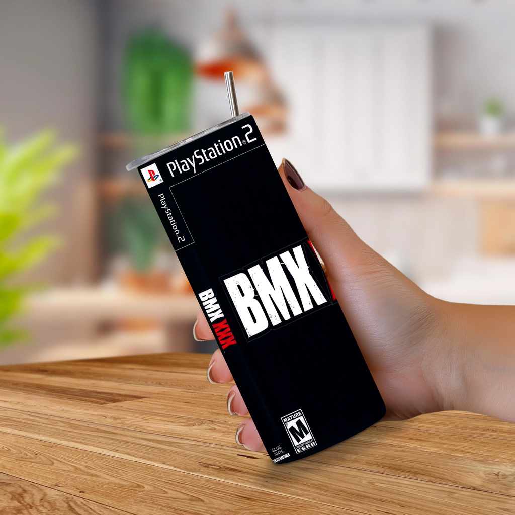 A hand holding a 20oz stainless steel tumbler BMX XXX PlayStation 2 game cover with a metal straw, set against a blurred kitchen background.