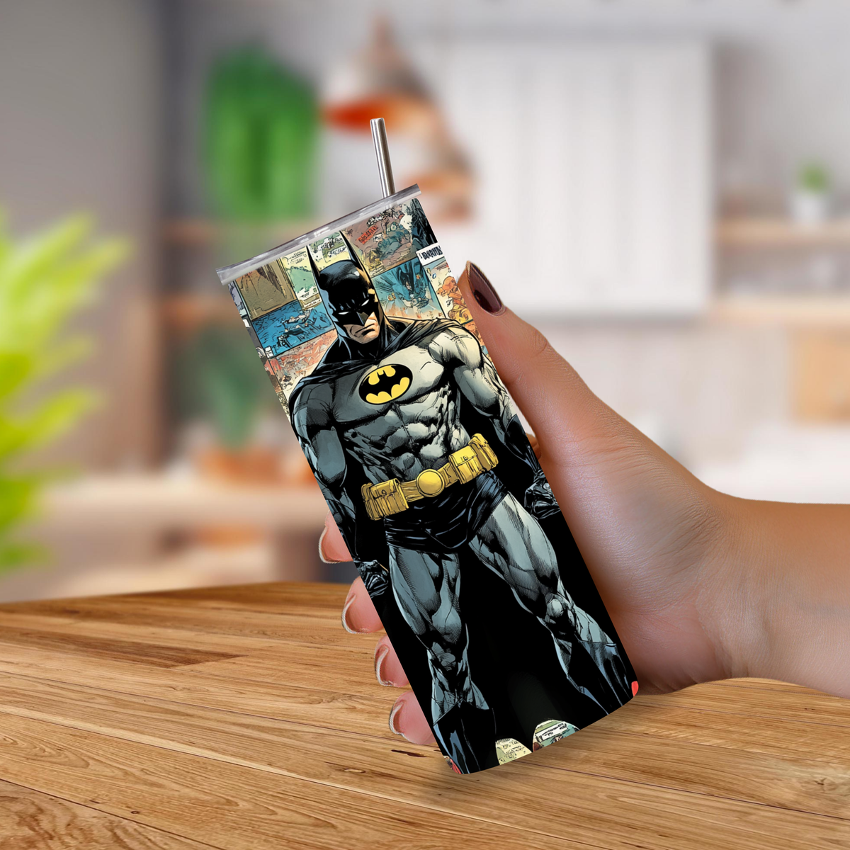A hand holding a slim stainless steel tumbler with a clear lid and straw, printed with a detailed Batman comic illustration, set against a blurred indoor background.
