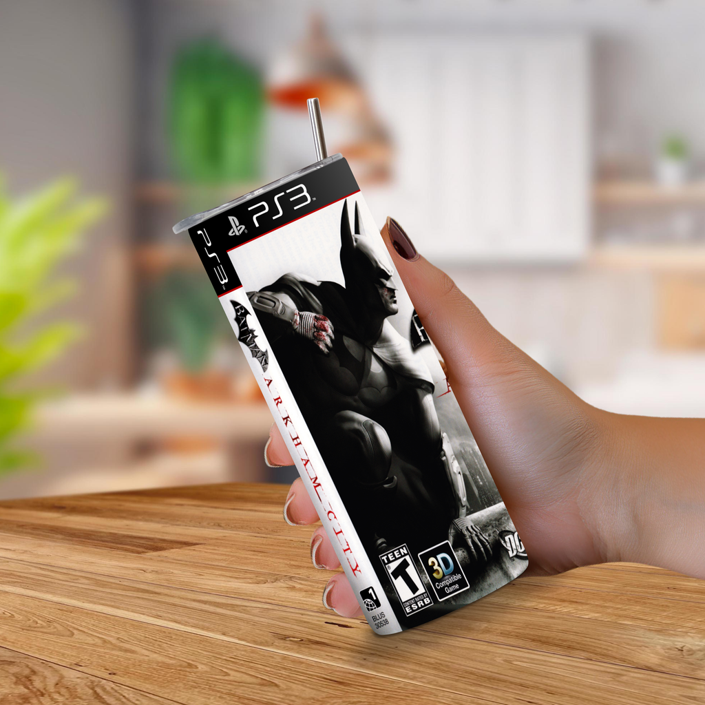A hand holding a 20oz tumbler with PS3 Batman: Arkham City design, featuring a printed game cover art and a metal straw, set against a blurred indoor background.