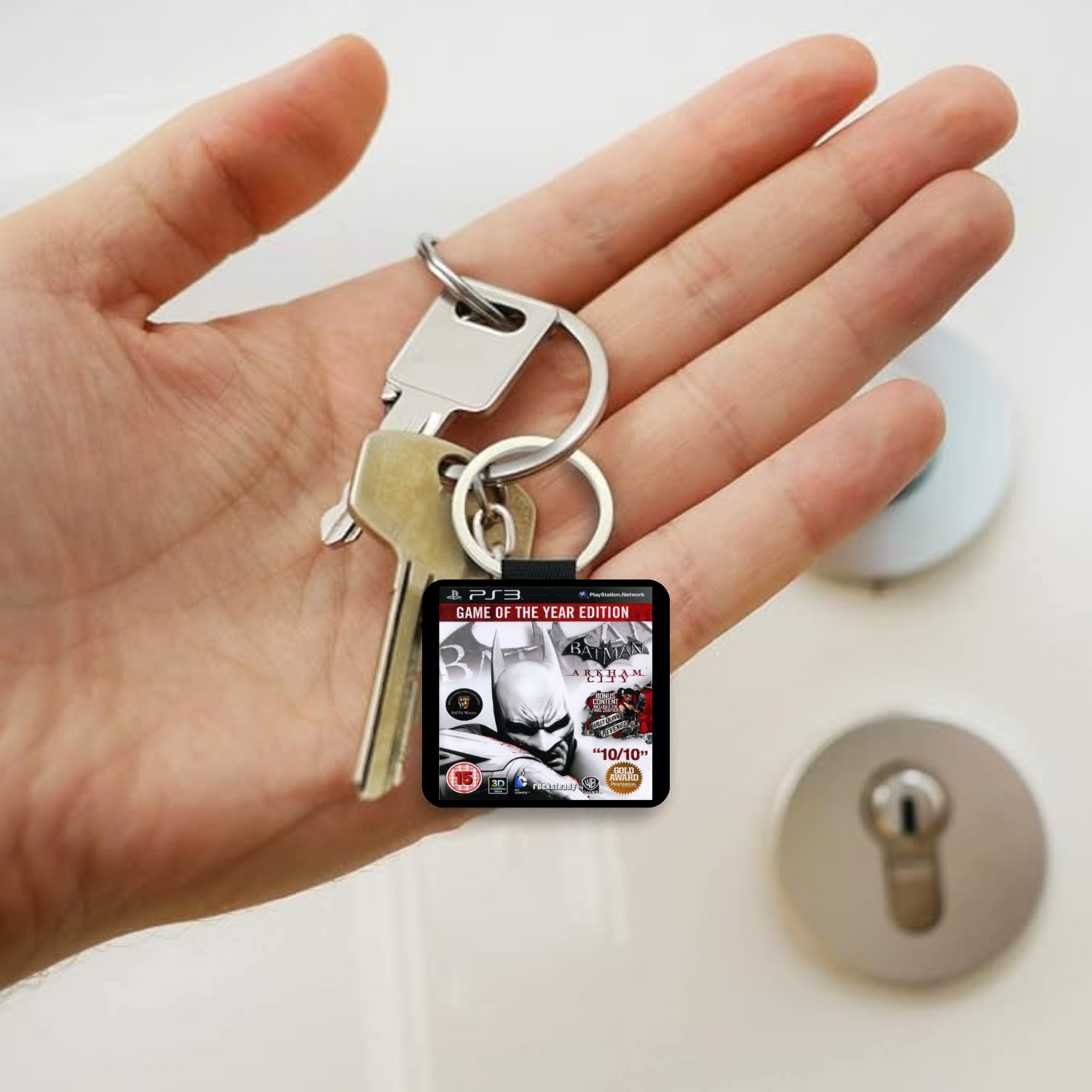 A hand holding a keyring with a small square PlayStation 3 Batman Arkham City 'Game of the Year Edition' keychain, with two metal keys attached. The background shows a white surface with a lock and lock cylinder blurred in the background.