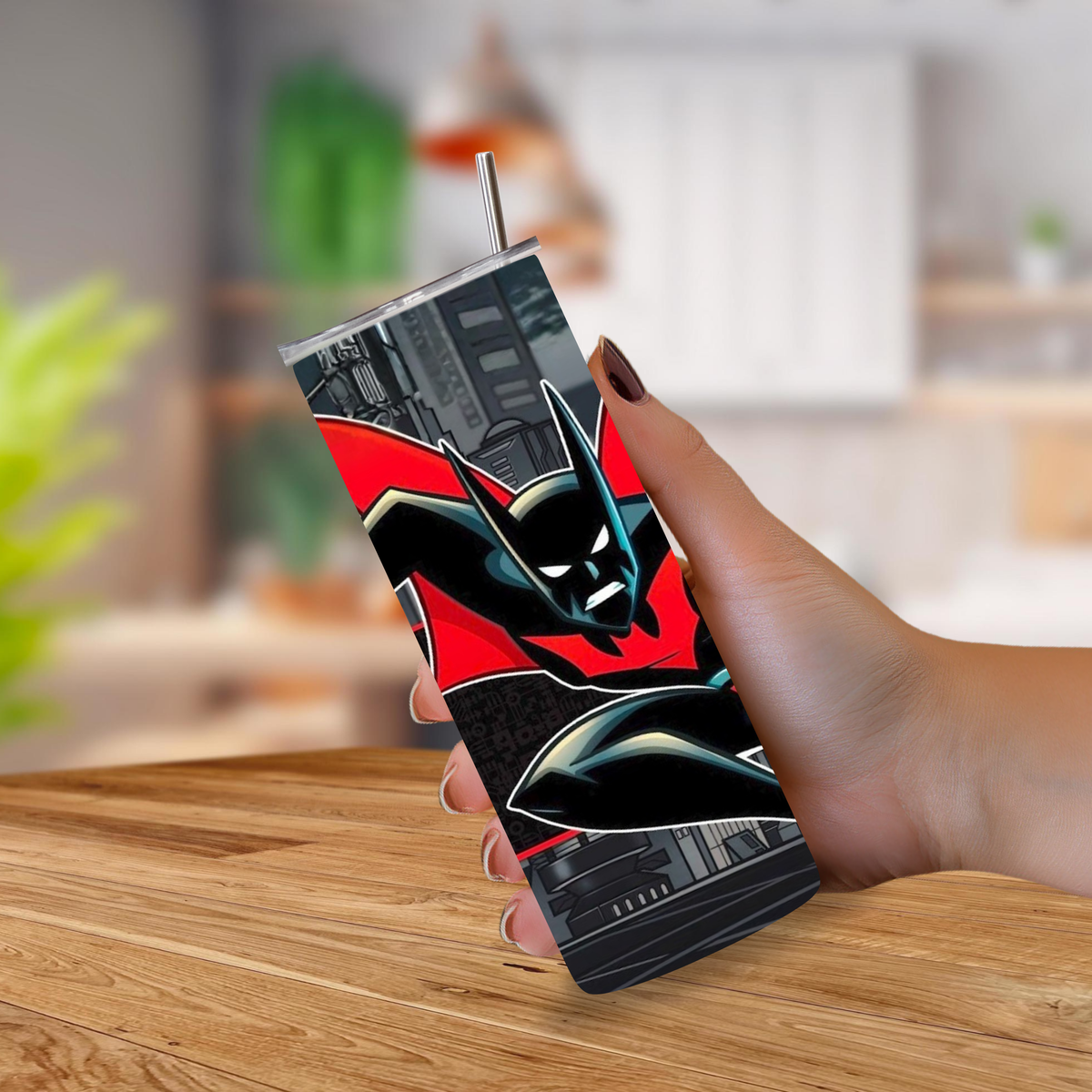 A hand holding a slim rectangular tumbler with a Batman graphic design and a metal straw, set against a blurred indoor background with a wooden table.