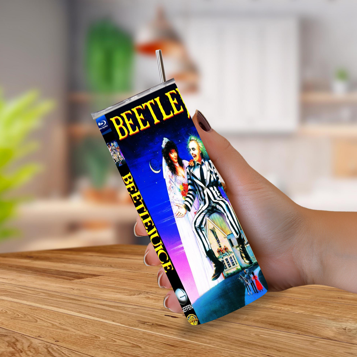 A hand holding a slim tumbler cup with a metal straw, printed with Beetlejuice movie artwork, against a blurred indoor background with a wooden table visible.