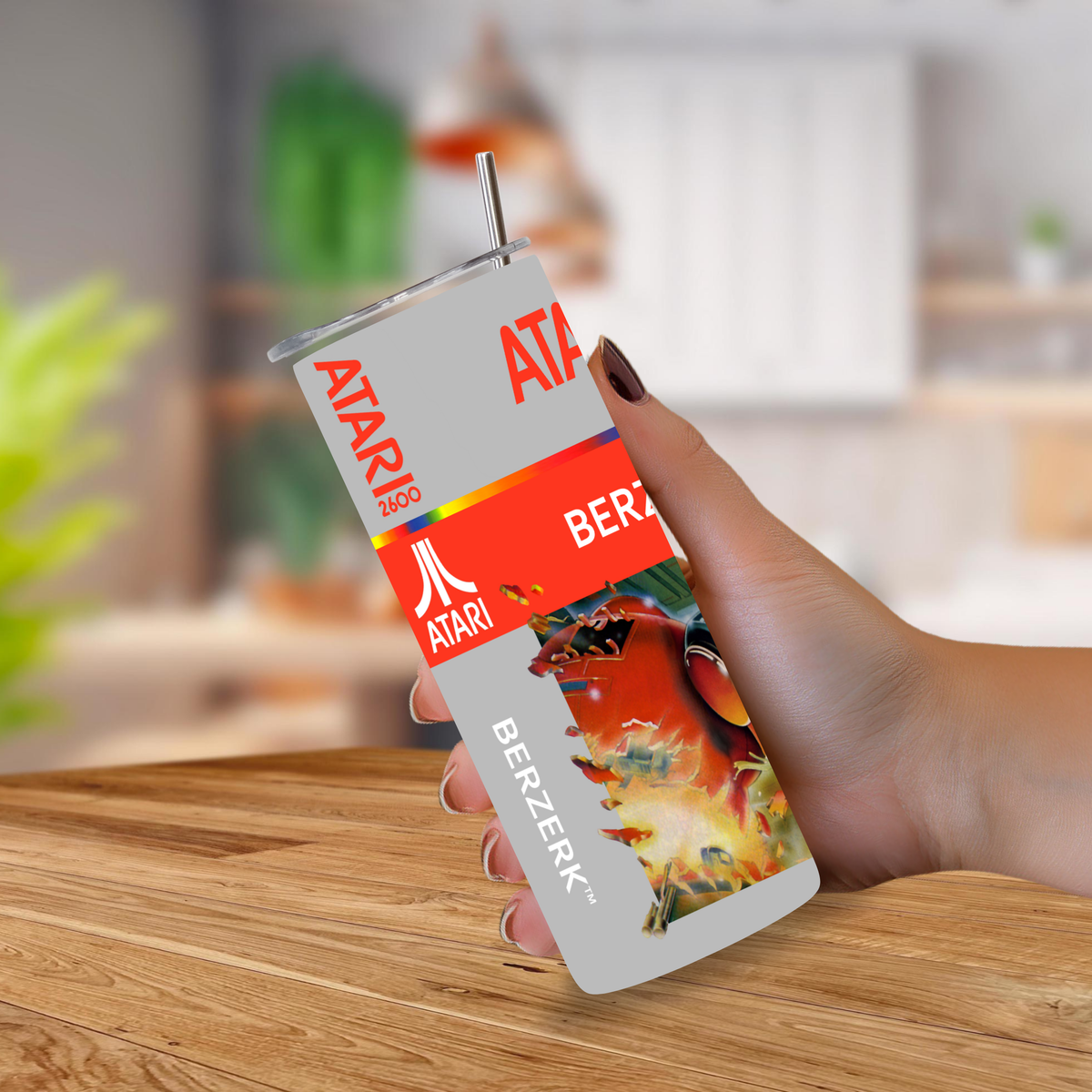 A hand holding a 20oz stainless steel tumbler with a metal straw, printed with retro Atari-themed graphics including the Atari logo, 'BERZERK' text, and a fiery red/orange graphic, against a blurred indoor background.
