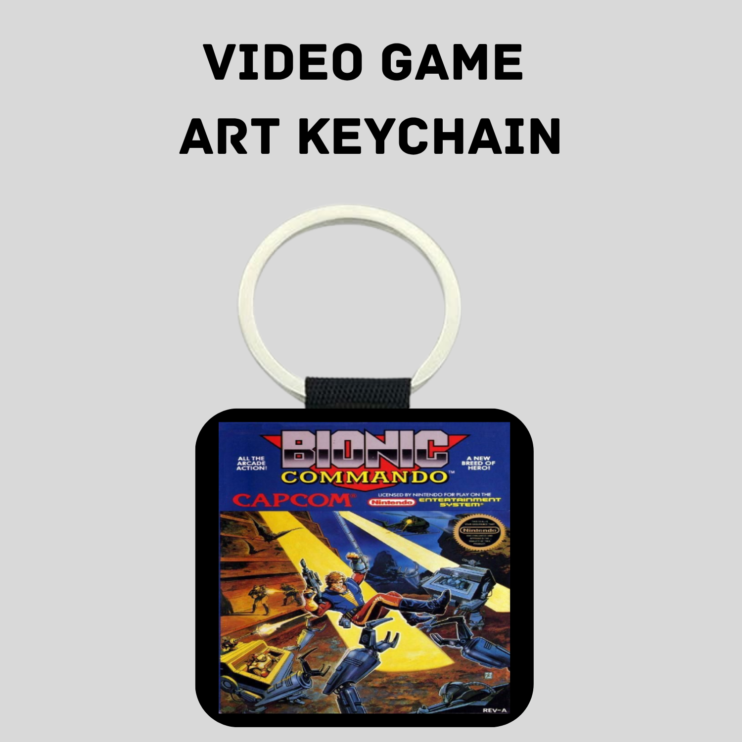 A square video game art keychain featuring Bionic Commando Capcom artwork, suspended from a white plastic ring with a black strap, set against a light gray background with the text 'VIDEO GAME ART KEYCHAIN' above it.