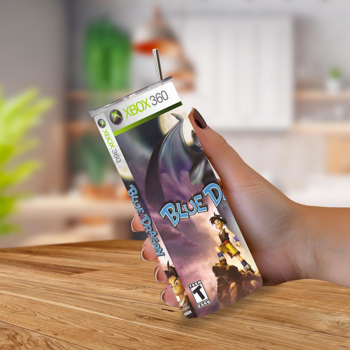 A hand holding a custom 20oz tumbler designed to look like an Xbox 360 Blue Dream game case, complete with a straw, set against a blurred kitchen background.
