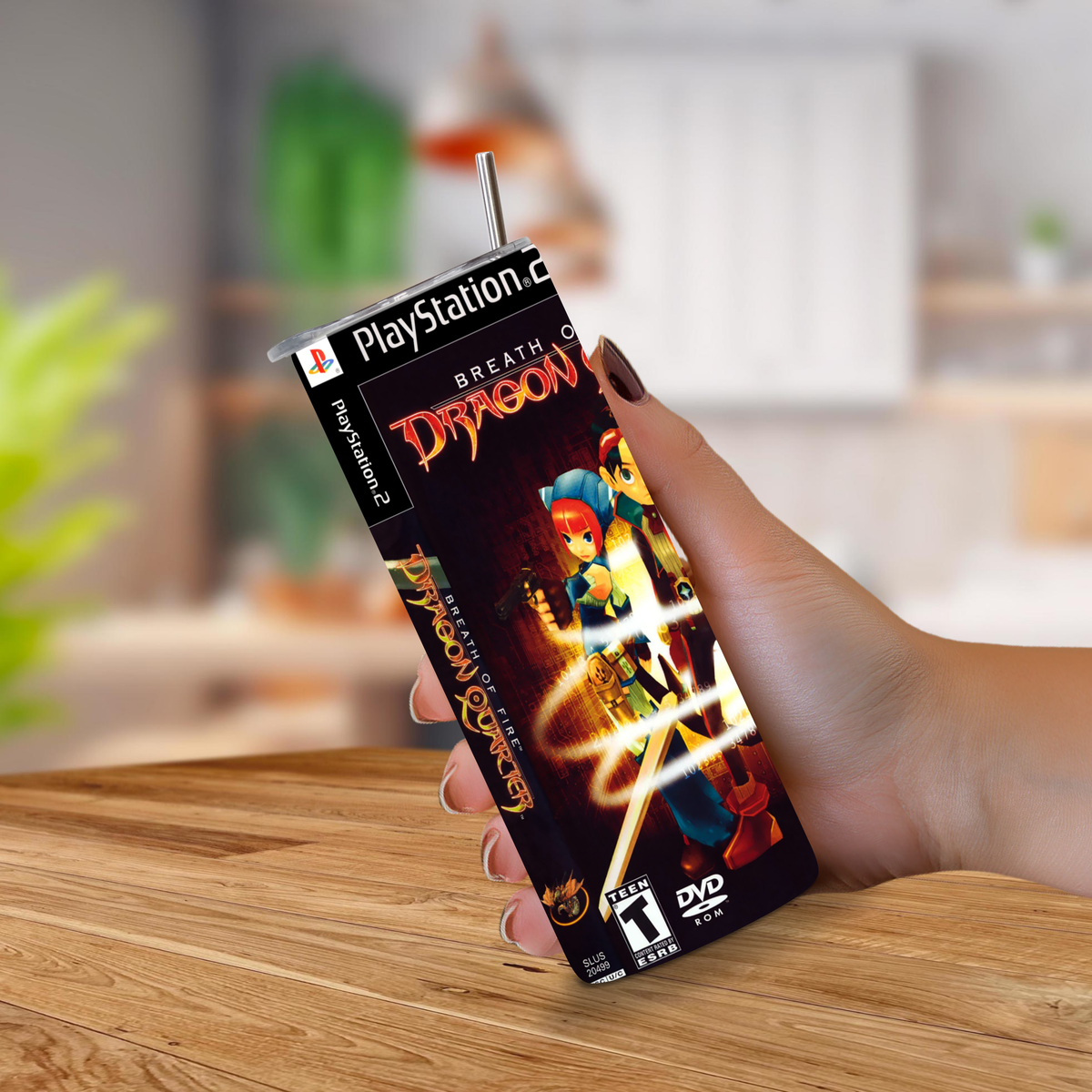 A hand holding a 20oz stainless steel tumbler, featuring Breath of Fire Dragon Quarter artwork, with a metal straw sticking out from the top, set against a blurred indoor background.