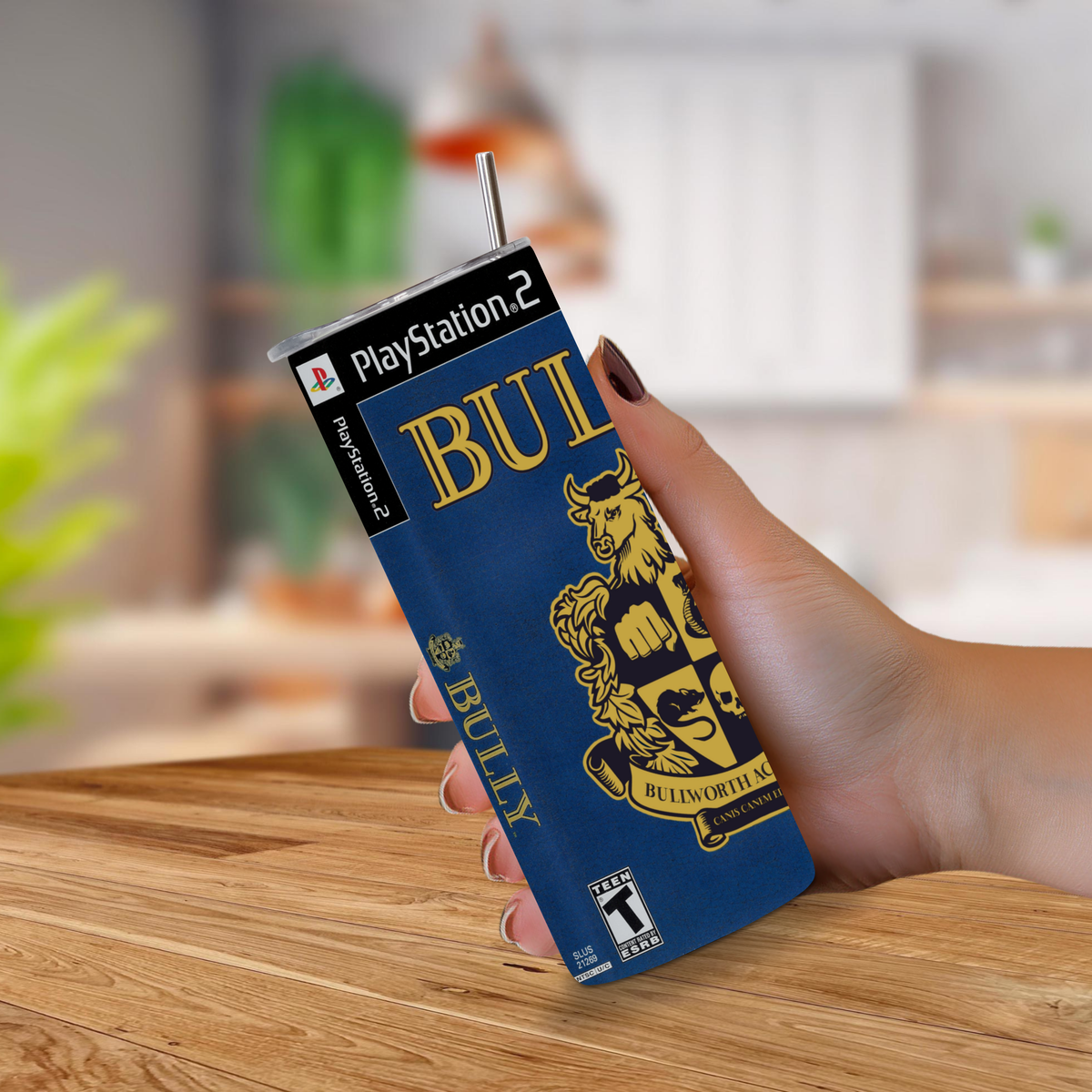A hand holding a PlayStation 2-themed j20oz tumbler with a straw, featuring a blue and gold 'BULLY' graphic and a blurred indoor kitchen background.