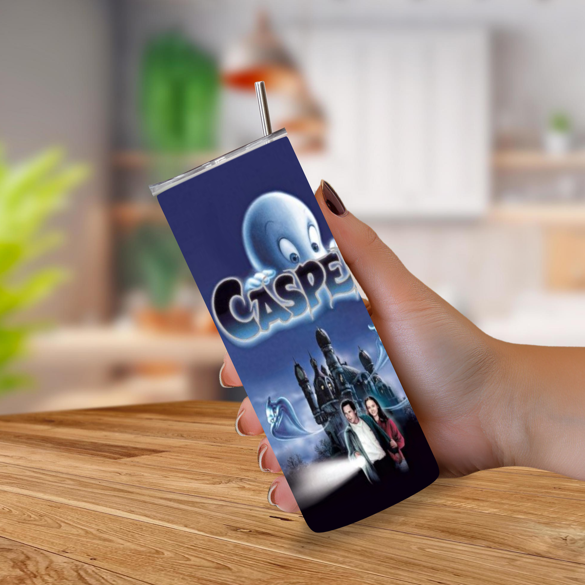 A hand holding a slim 20oz tumbler with a metal straw, featuring a printed Casper movie graphic on a dark blue background, set against a blurred indoor background with a wooden table.