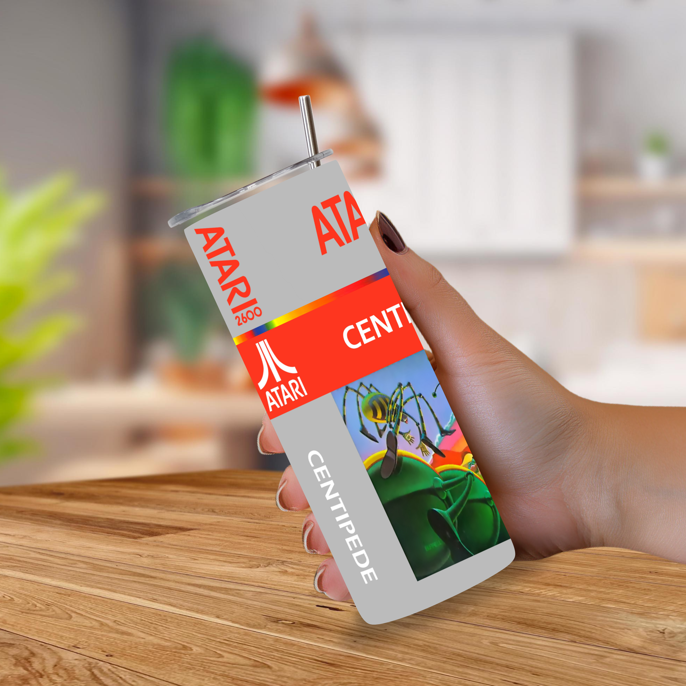 A hand holds a tall, slim Atari Centipede cup against a blurred indoor background. The cup is light gray with red Atari branding, a bright orange band, and a colorful graphic of a green centipede on a blue background.