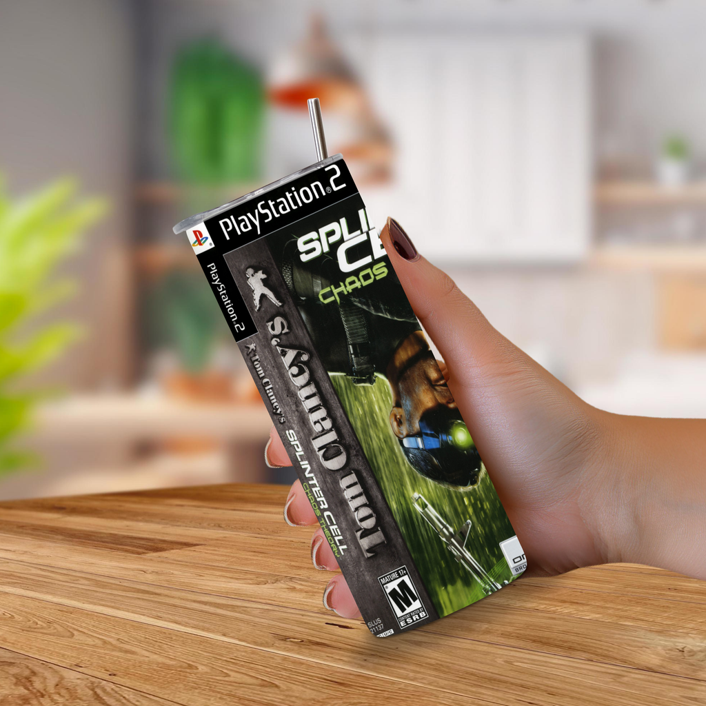A hand holds a PlayStation 2 20oz stainless steel tumbler, featuring the Splinter Cell Chaos Theory game art, against a blurred indoor background with a wooden table.