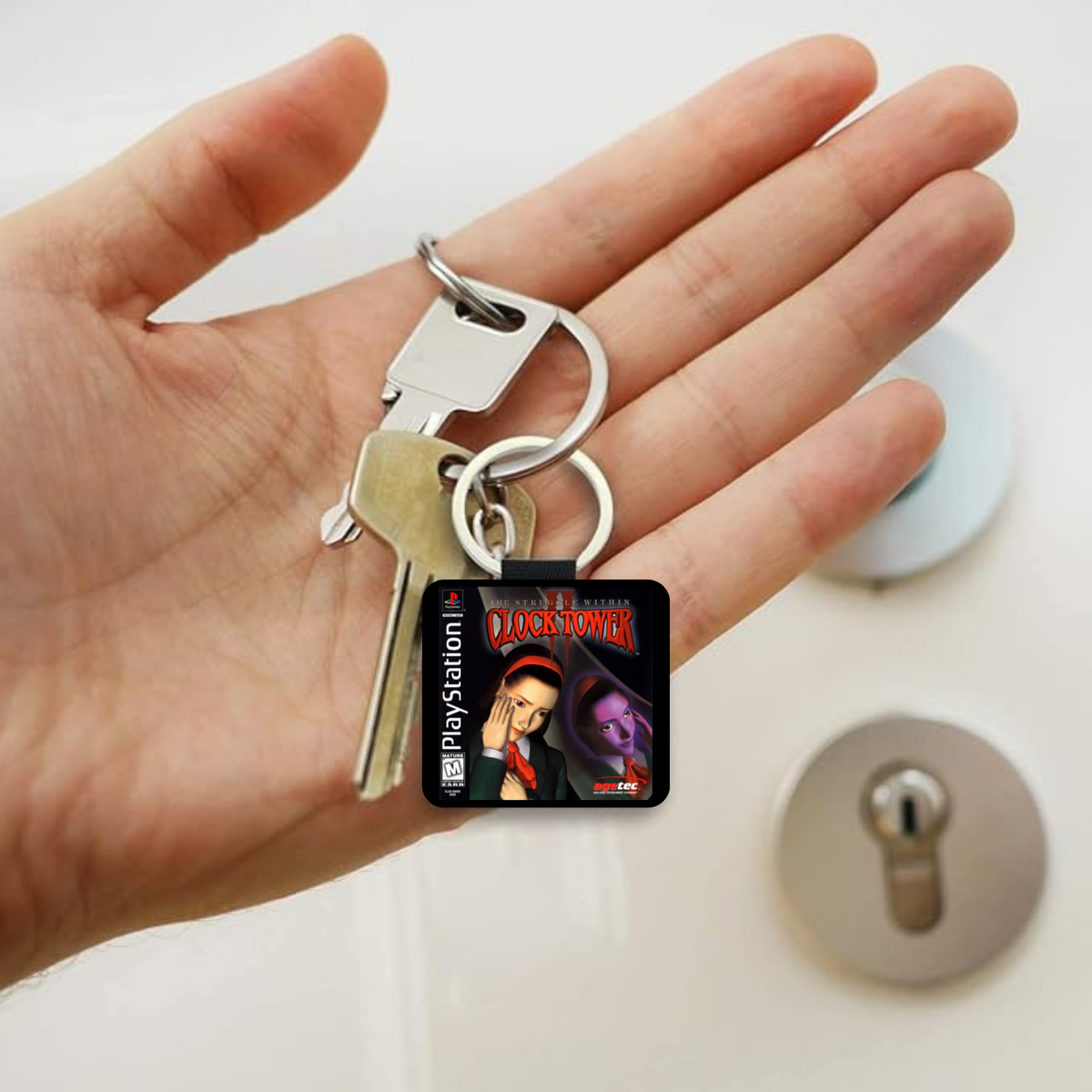 A hand holding a silver keyring with a square PlayStation-themed keychain featuring a black background and colorful Clock Tower II character artwork, with two metal keys attached.