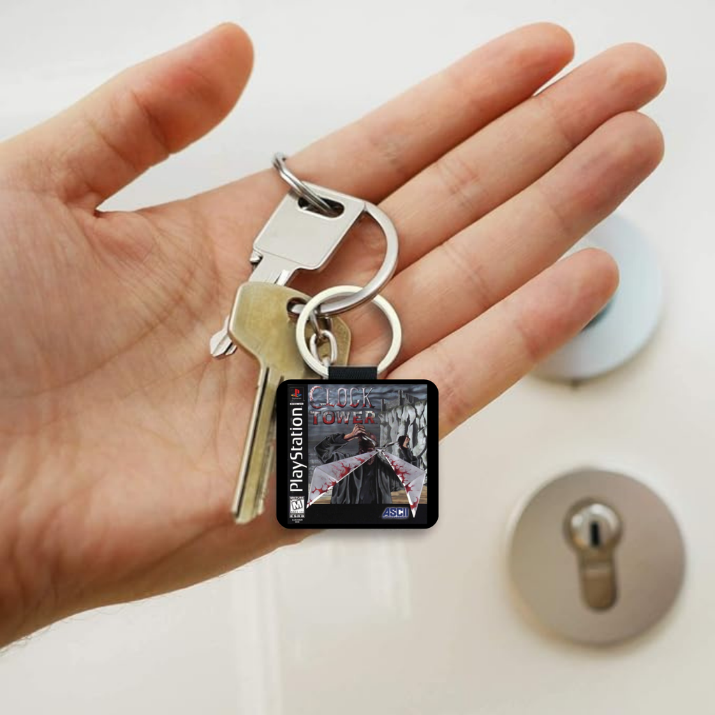 A hand holding a PlayStation keychain with two keys and a small square charm displaying the PlayStation logo and game artwork from Clock Tower, with a lock and lockout panel in the blurred background.