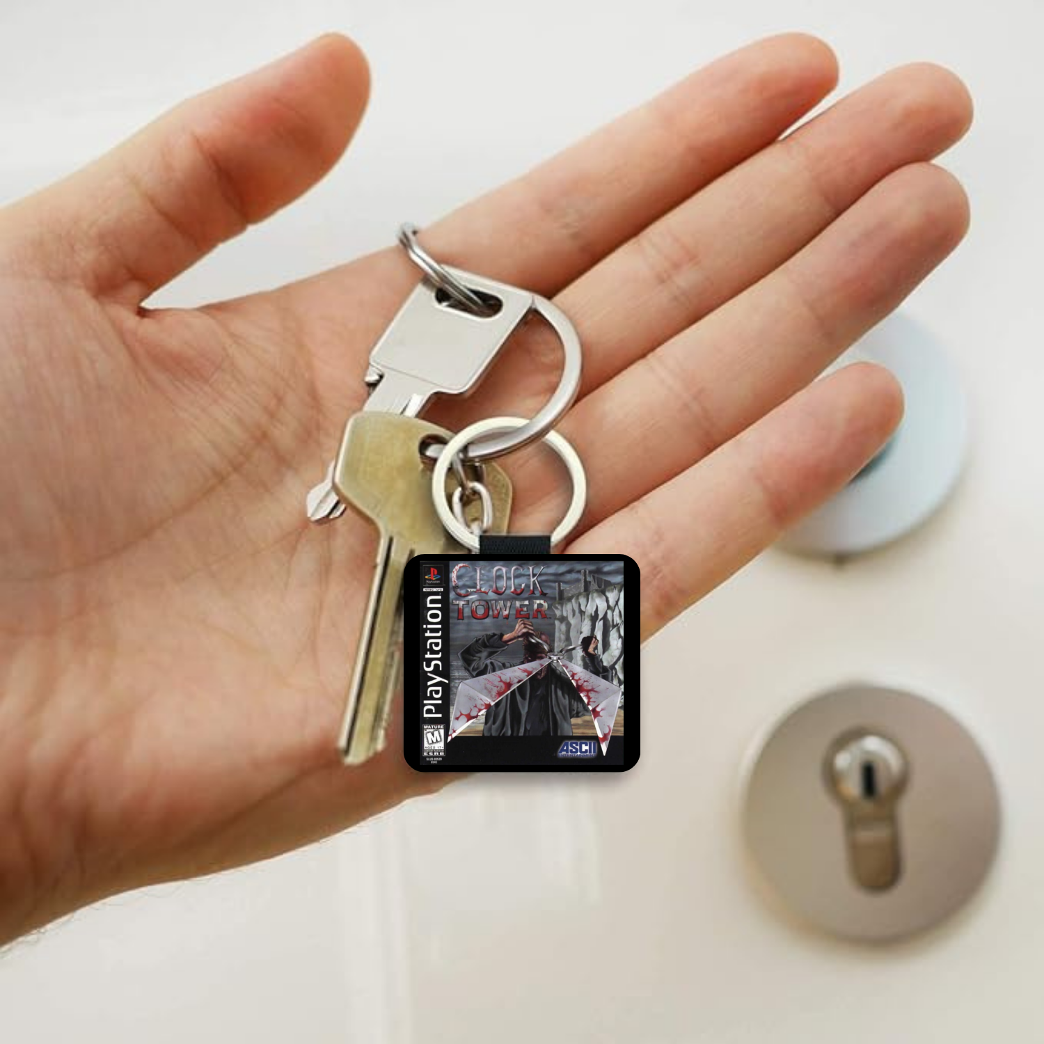 A hand holding a PlayStation keychain with two keys and a small square charm displaying the PlayStation logo and game artwork from Clock Tower, with a lock and lockout panel in the blurred background.