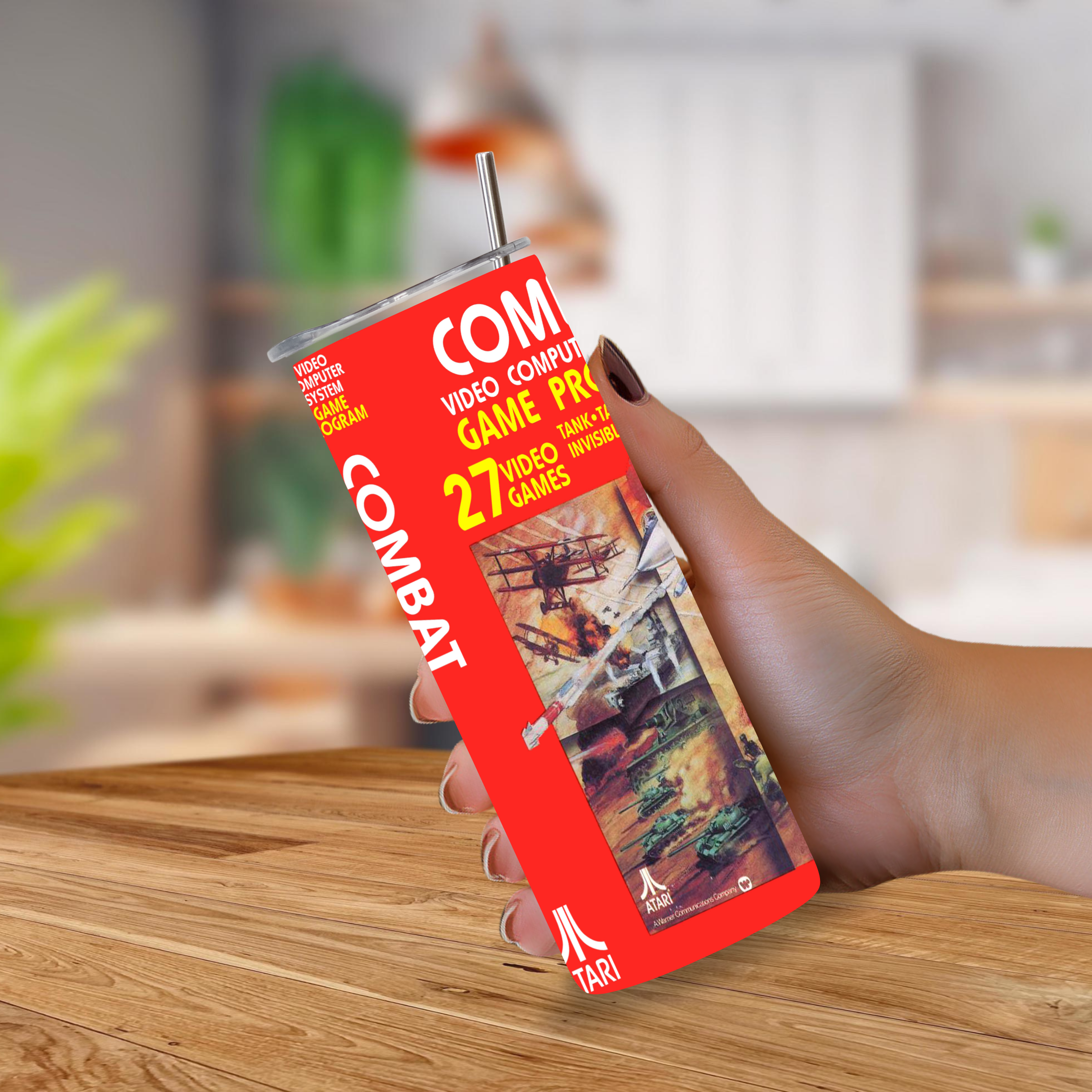 A hand holding a custom-designed 20oz tumbler cup with a metal straw. The cup is designed to look like a vintage Atari game case, featuring a bright red background, white text reading 'COMBAT'. 