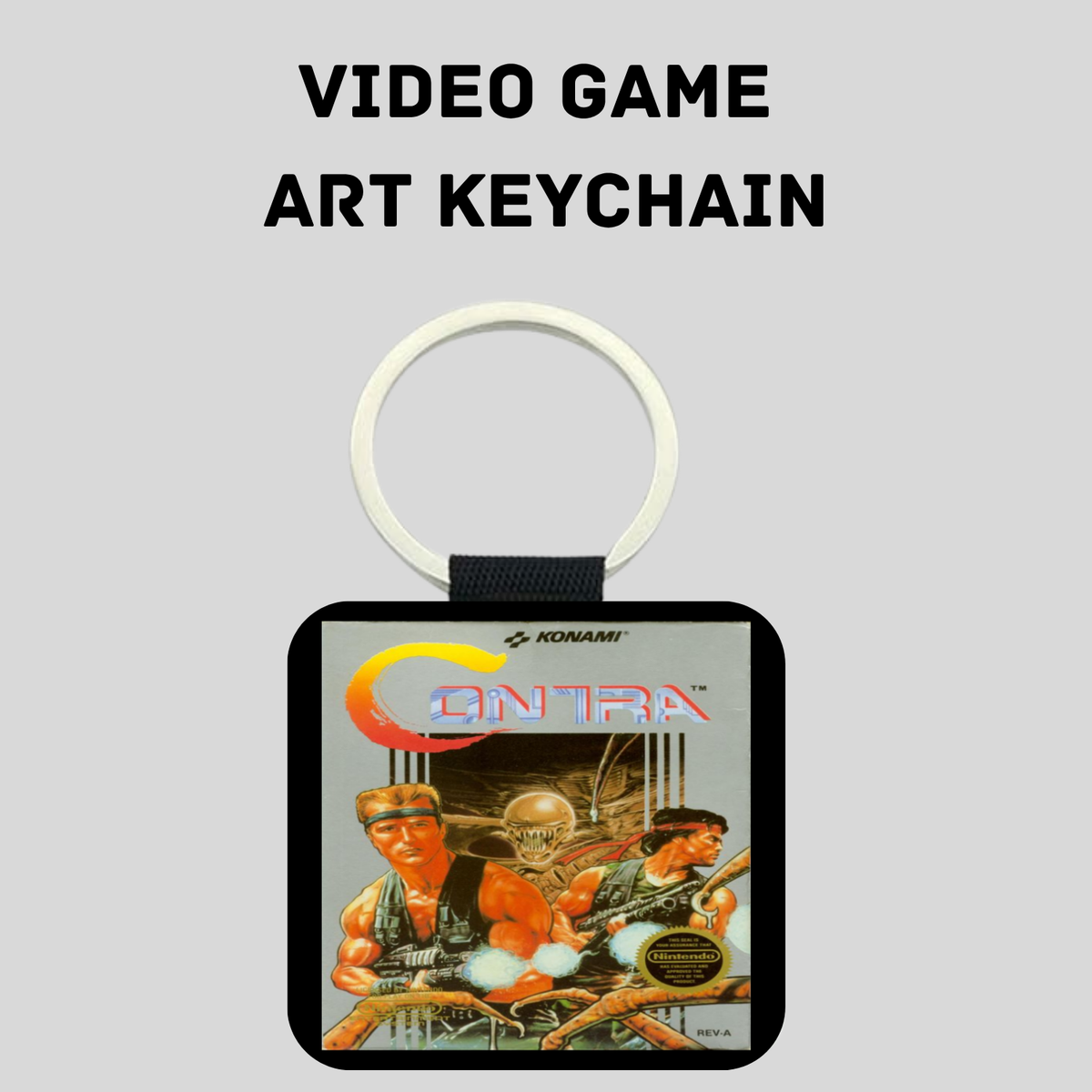A square keychain featuring retro Contra video game artwork, with a white split ring and black strap, shown on a light gray background with the text 'VIDEO GAME ART KEYCHAIN' above it.