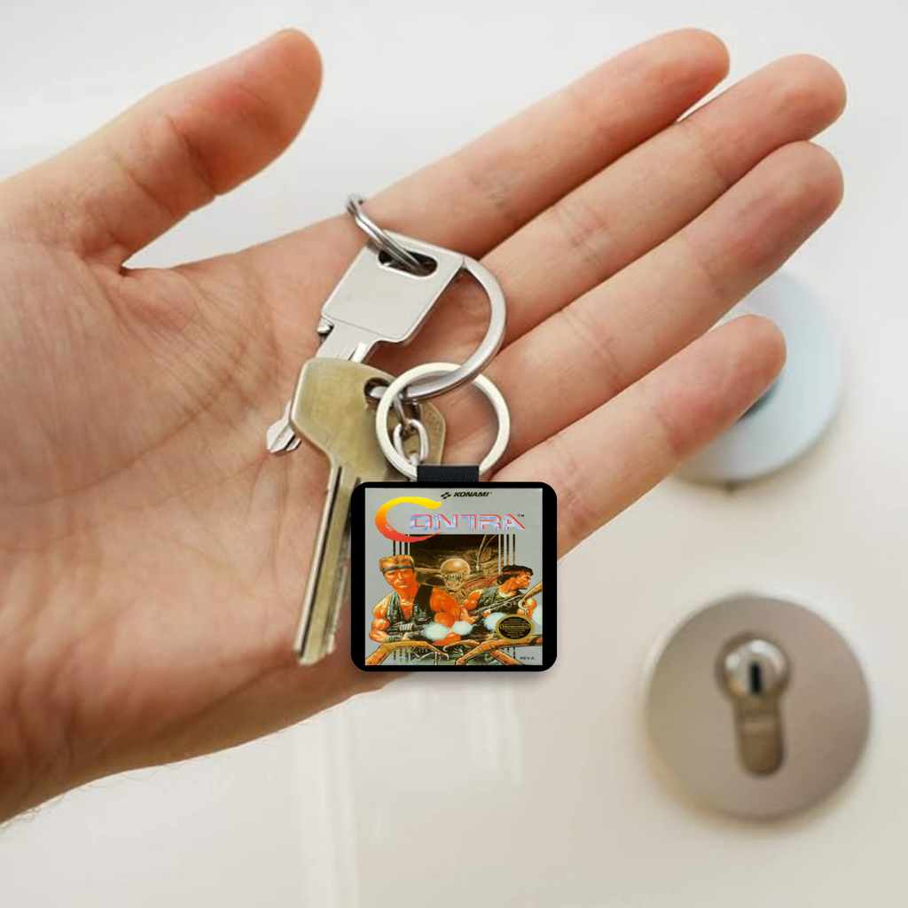 A hand holding a silver keyring with two keys and a square keychain featuring the NES Contra video game.