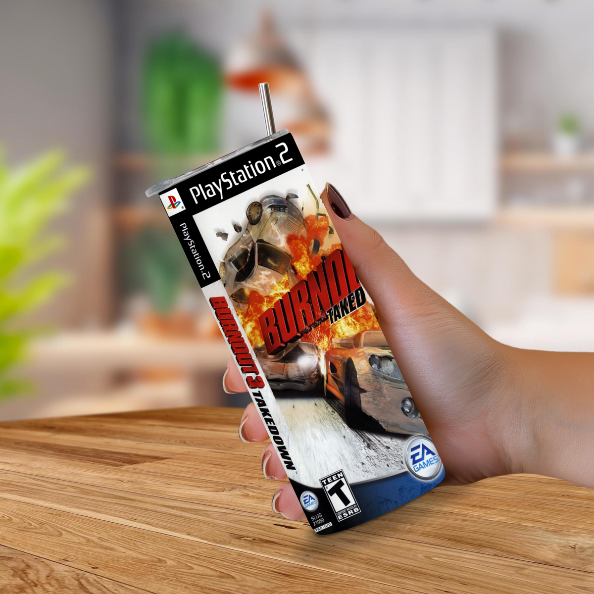 A hand holding a novelty PlayStation 2 game case designed to look like a 20oz stainless steel tumbler, featuring the game cover art for 'Burnout 3 Takedown' with a metal straw inserted at the top, set against a blurred indoor background.