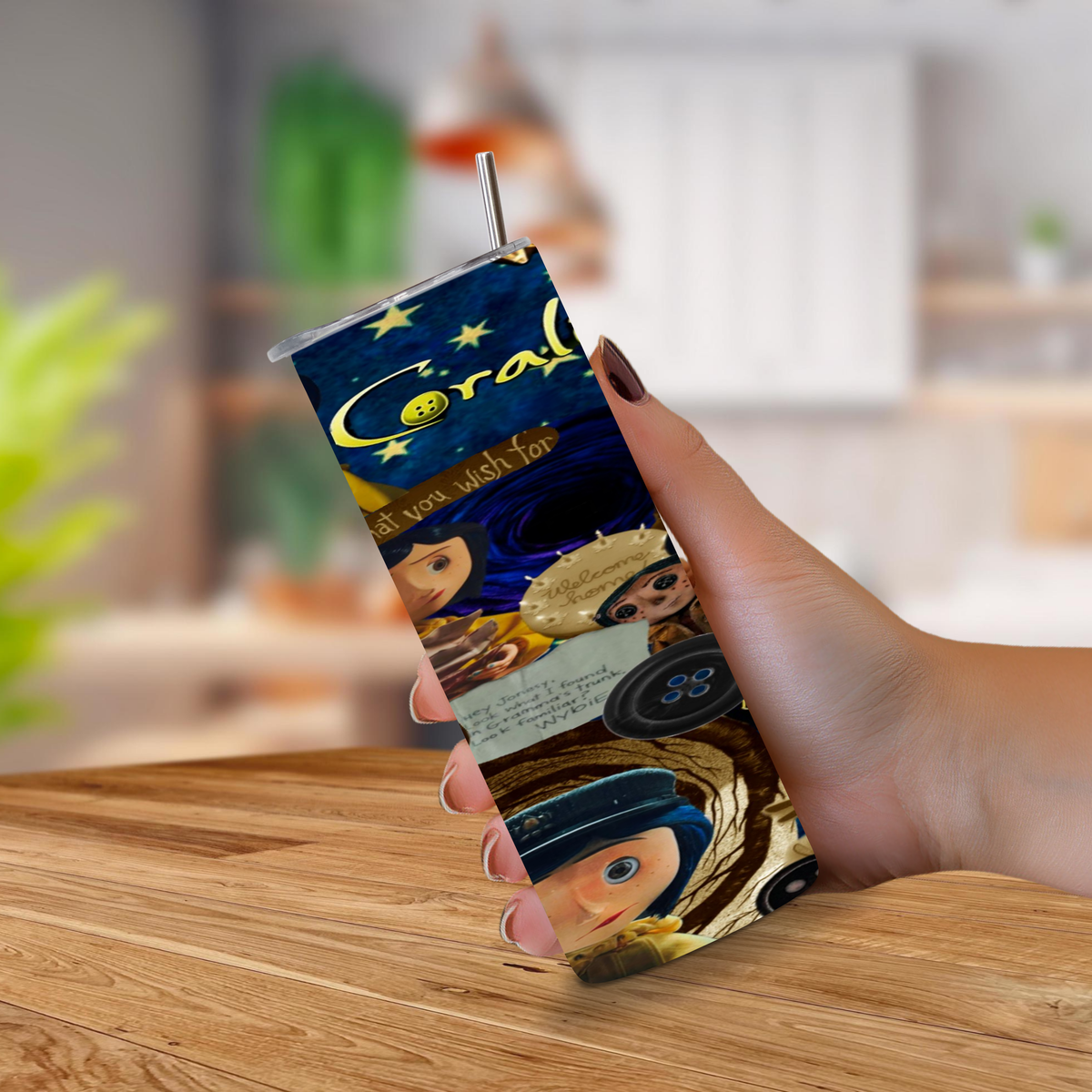 A hand holding a clear plastic tumbler with a printed Coraline theme and with a metal straw, set against a blurred kitchen background.