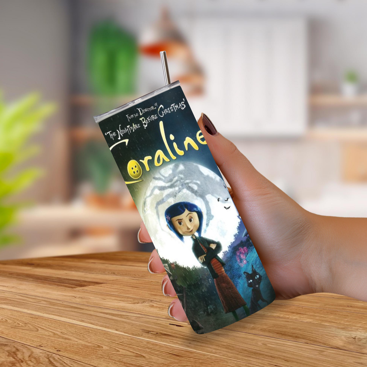 A hand holding a stainless steel tumbler with a straw, printed with the 'The Nightmare Before Christmas' Coraline character artwork, set against a blurred kitchen background.
