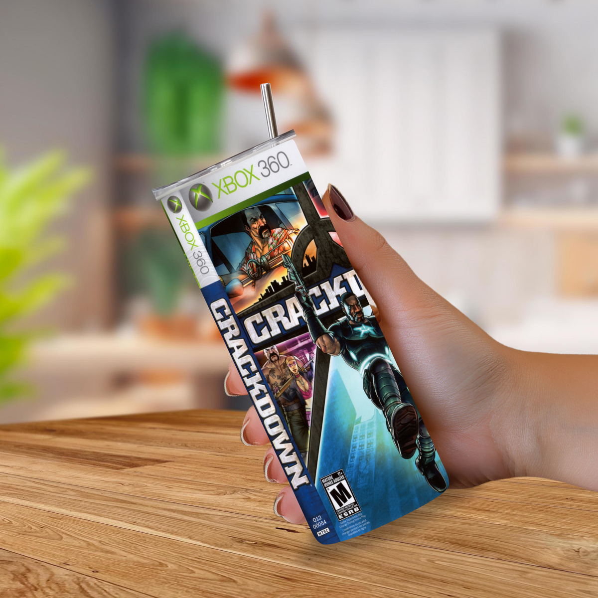 A hand holding a Crankdown XBOX 360 themed 20oz tumbler with a metal straw, set against a blurred indoor background.