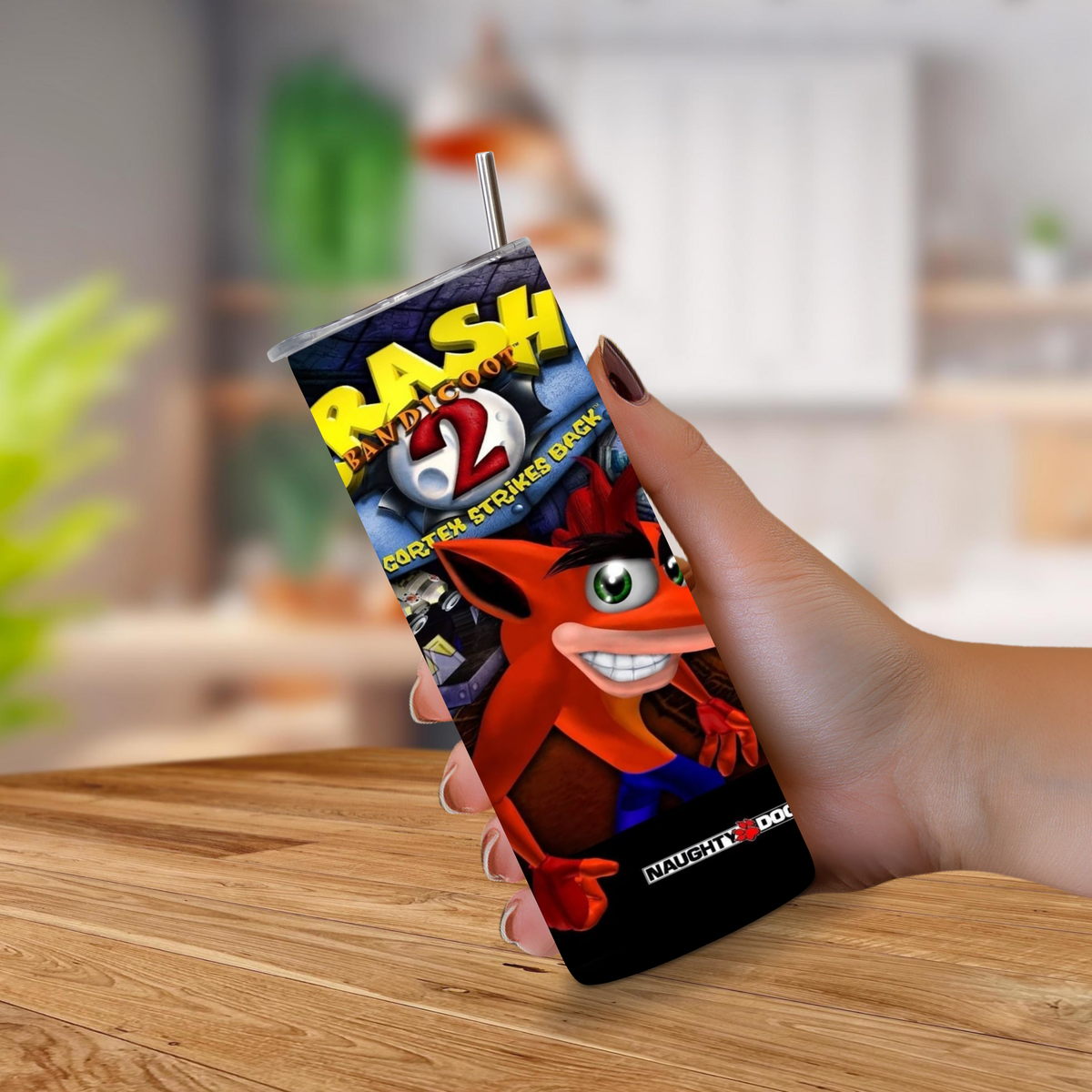A hand holding a Crash Bandicoot 2 themed insulated tumbler with a straw, set against a blurred kitchen background.