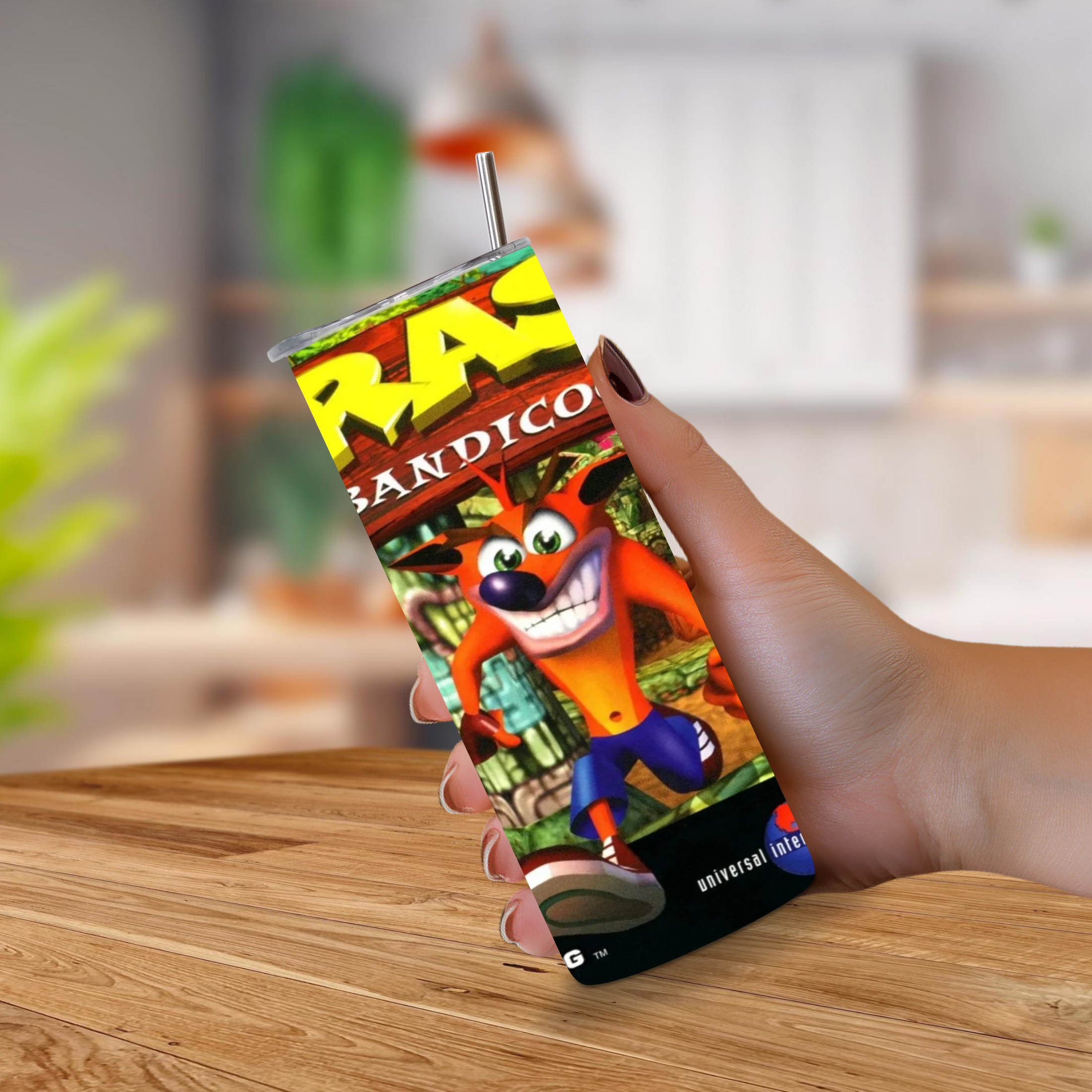 A hand holding a 20oz stainless steel tumbler with a metal straw, featuring a colorful Crash Bandicoot video game graphic on the front, set against a blurred indoor background.