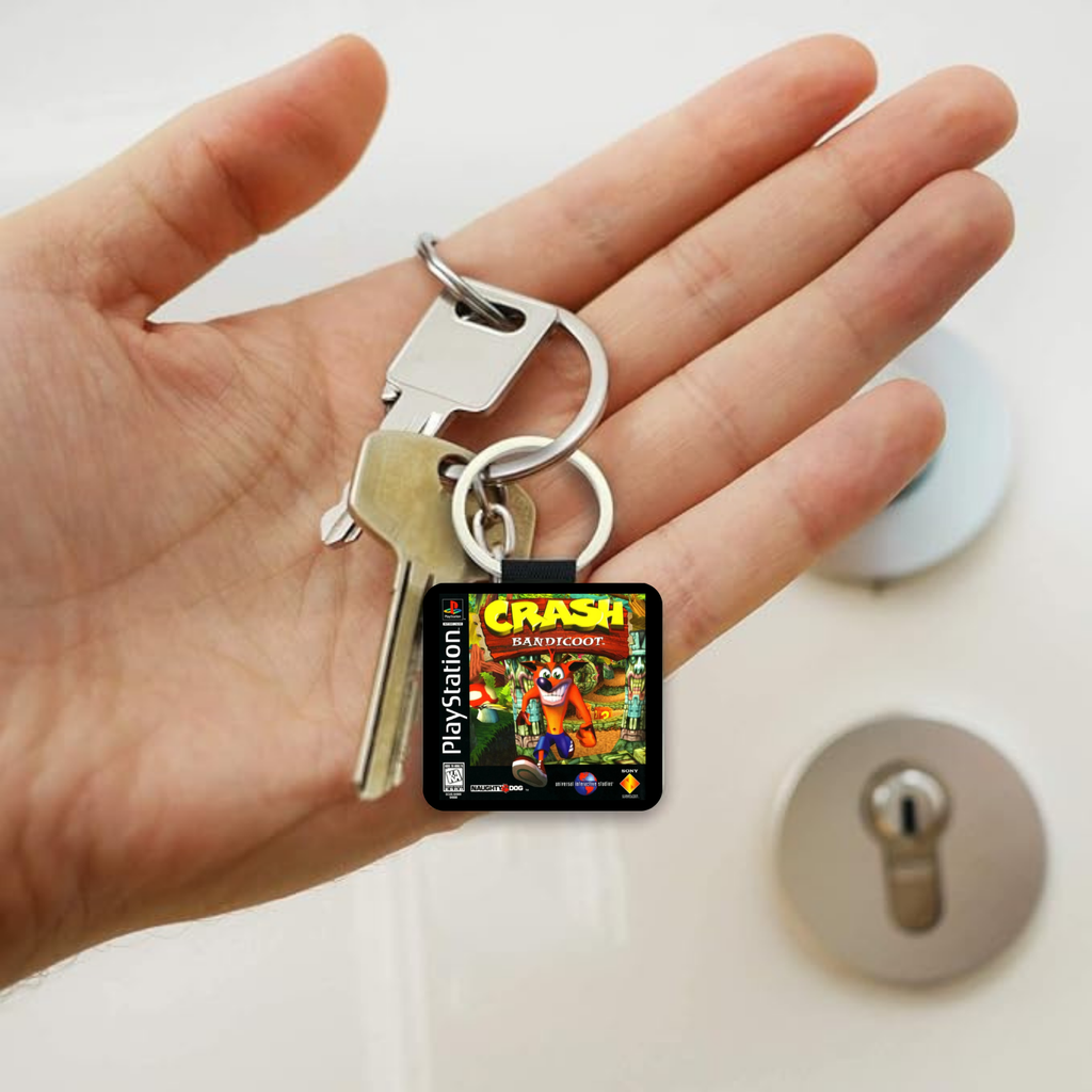 A hand holding a keyring with two keys and a small square Crash Bandicoot PlayStation keychain charm, with a lock and lockout plate blurred in the background.