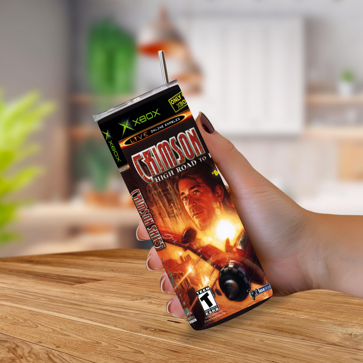 A hand holding a custom-printed Xbox-branded tumbler cup featuring the video game cover art for 'Crimson Skies: High Road to Revenge'. The cup has a clear plastic lid with a metal straw and is shown against a blurred indoor background with a wooden table.