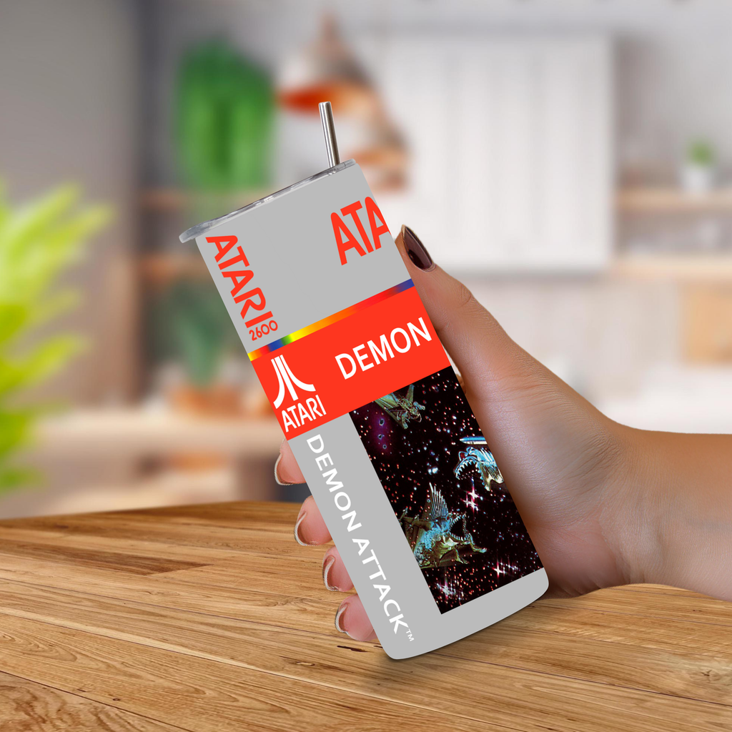 A hand holding a retro Atari 'Demon Attack' themed tumbler cup with a straw, set against a blurred kitchen background.
