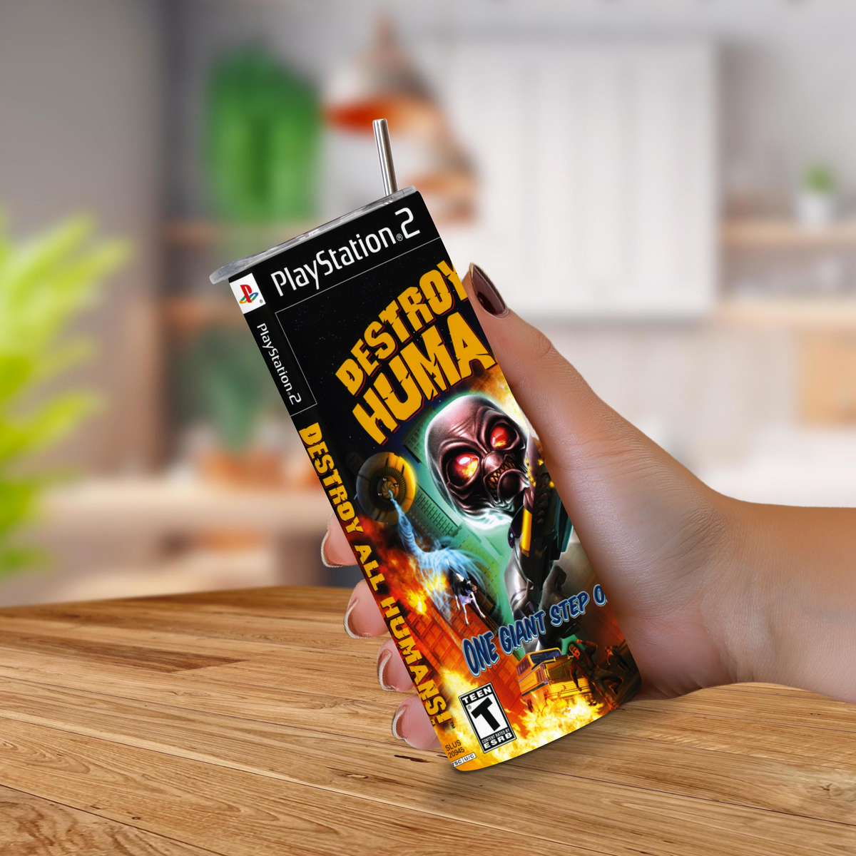 A hand holding a PlayStation 2-themed tumbler cup with a straw, featuring the 'Destroy All Humans' video game cover art, set against a blurred kitchen background.