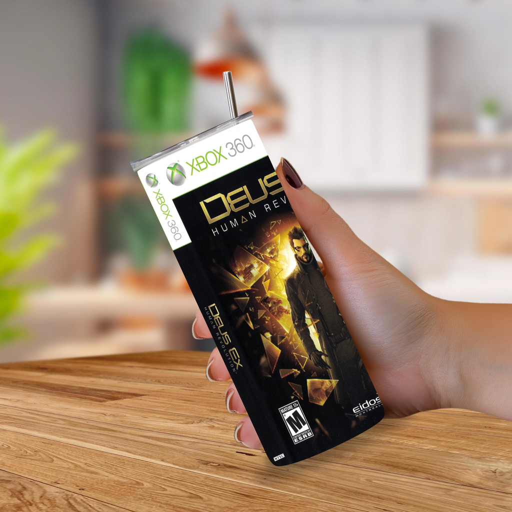 A hand holding a tumbler-shaped novelty drinkware item featuring the Xbox 360 and Deus Ex Human Revolution box art, with a straw inserted at the top, set against a blurred indoor background.