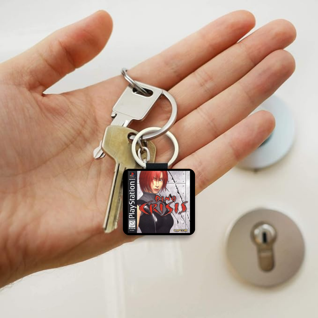 A hand holding a silver keyring with two keys and a square PlayStation-themed keychain featuring the character from Dino Crisis.