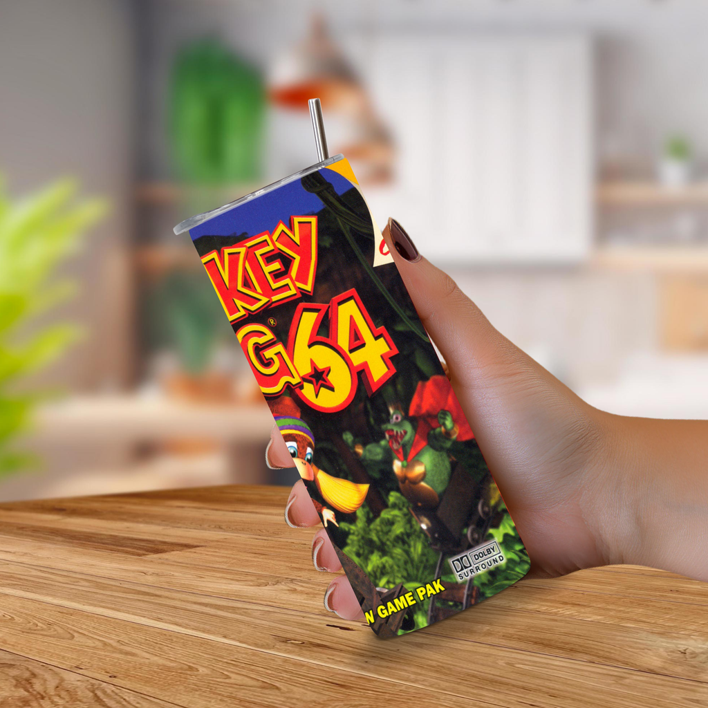A hand holding a 20oz stainless steel Donkey Kong 64 tumbler with a straw, set against a blurred indoor background on a wooden table.
