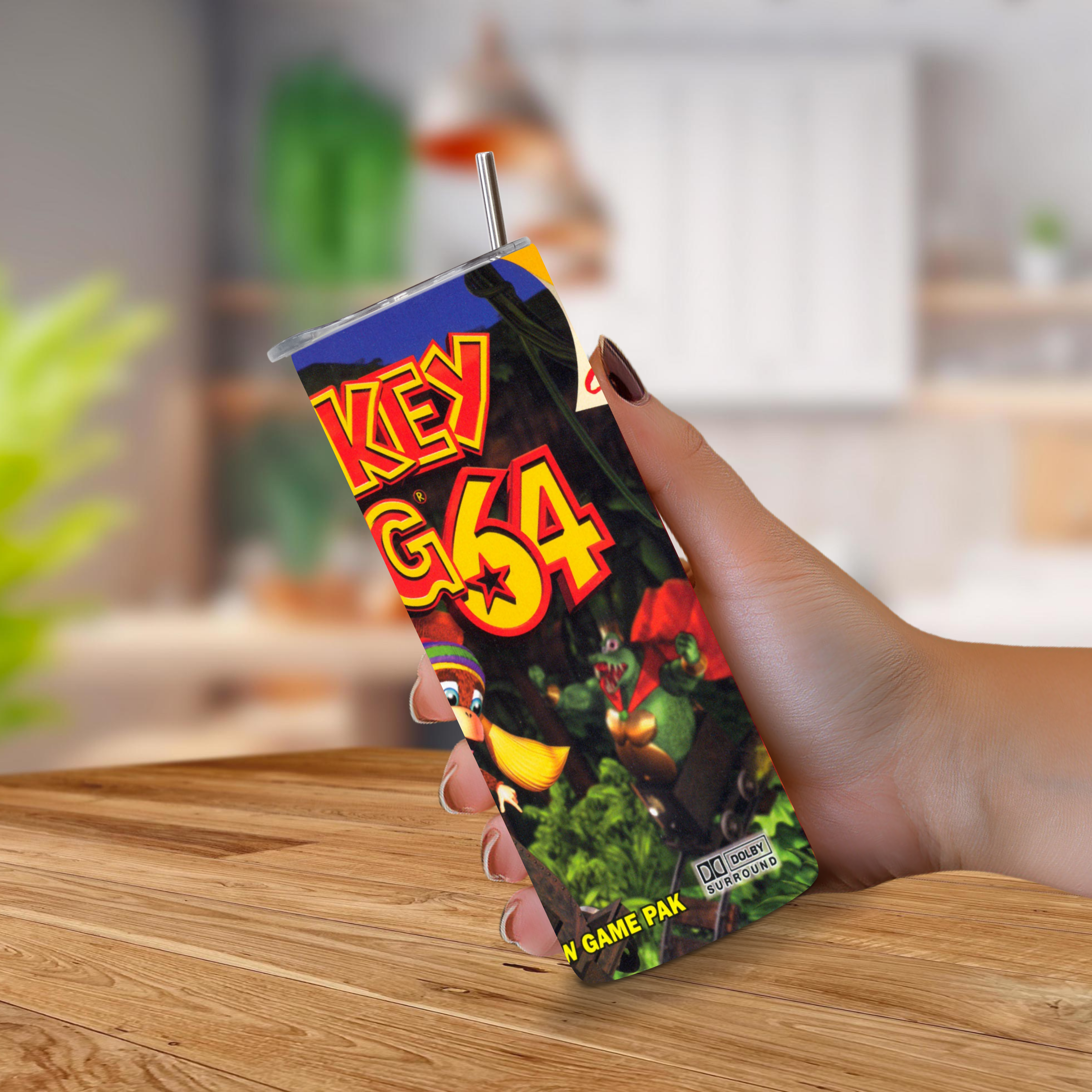 A hand holding a 20oz stainless steel Donkey Kong 64 tumbler with a straw, set against a blurred indoor background on a wooden table.