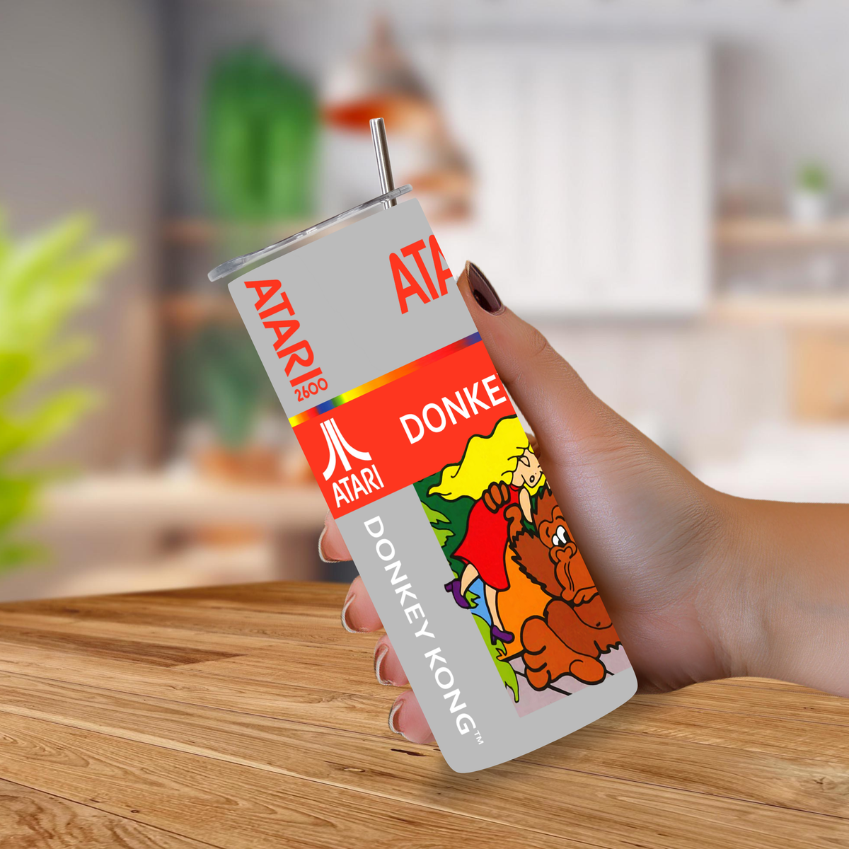 A hand holding a 20oz stainless steel tumbler with a metal straw, printed with Atari and Donkey Kong graphics, against a blurred indoor background.