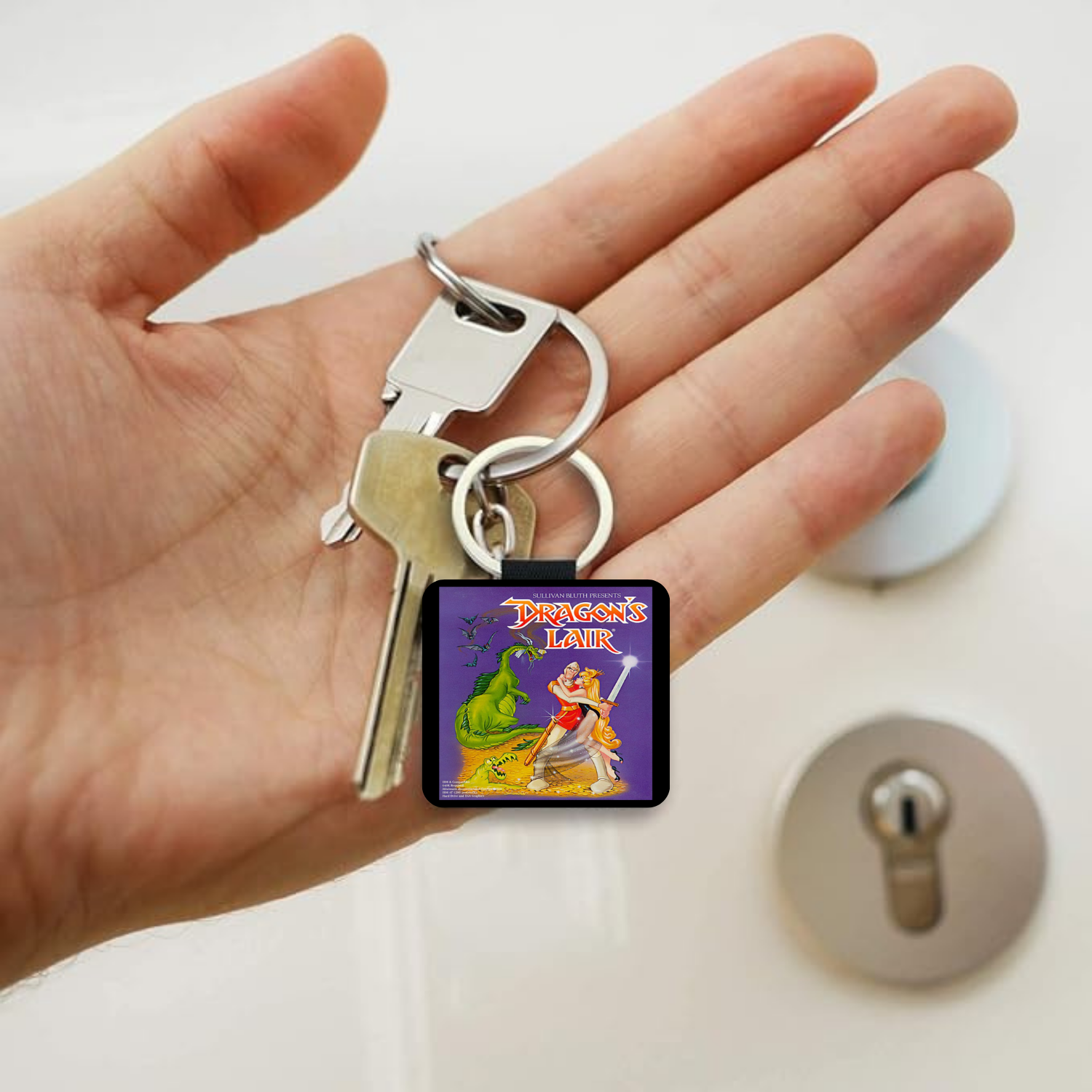 A hand holding a silver keyring with a square charm featuring a colorful cartoon illustration from the video game Dragon's Lair. 