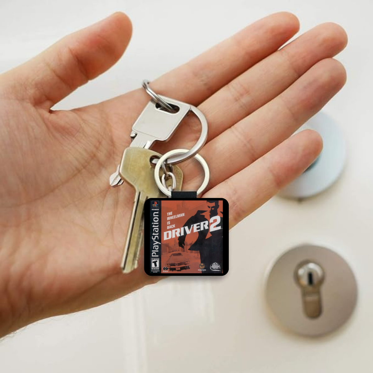 A hand holding a PlayStation Driver 2 themed keychain with a square black and red game cover charm, alongside two metal keys, against a blurred background of a lock and white surface.