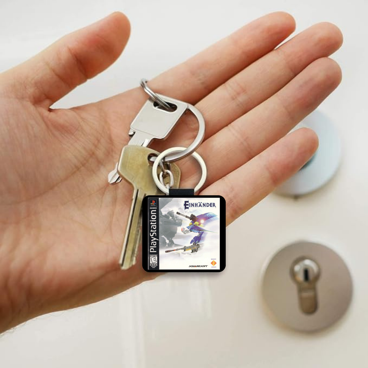 A hand holding a keyring with a small PlayStation game case charm featuring the Einhander game cover, alongside two metal keys, against a light background with a lock visible in the distance.