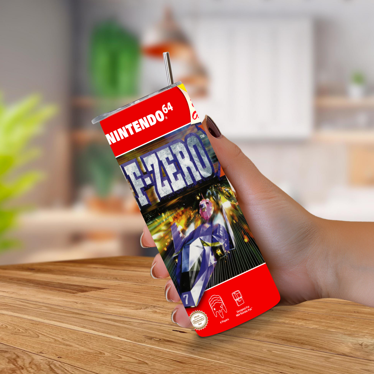 A hand holding a Nintendo 64 F-Zero themed 20oz tumbler with a straw, set against a blurred indoor background and wooden tabletop.