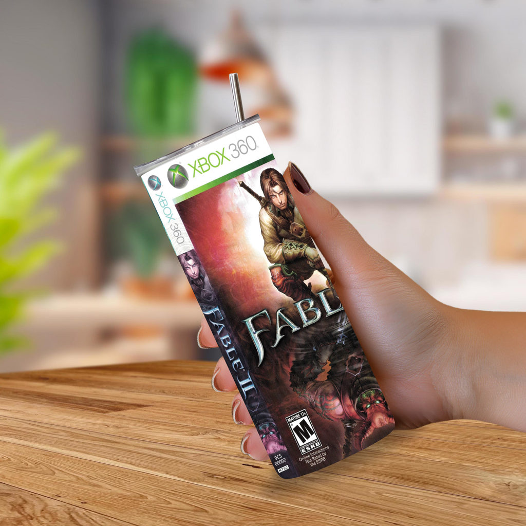A hand holding a custom 20oz tumbler designed to look like an Xbox 360 game case for the game Fable, with a metal straw inserted at the top, set against a blurred kitchen background.
