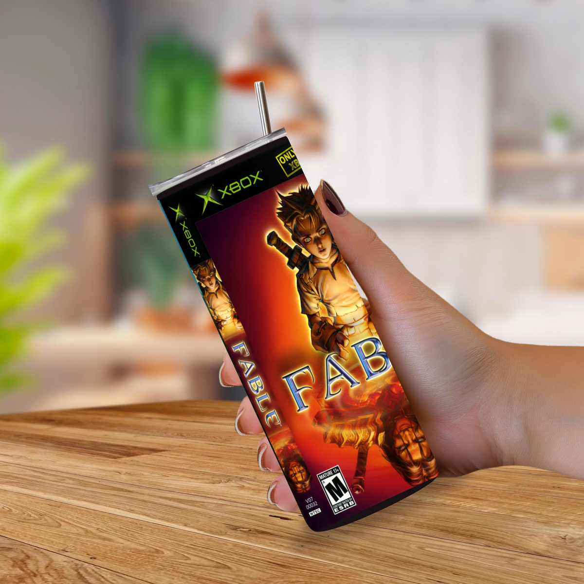 A hand holding a custom Xbox-branded tumbler cup with a straw, featuring a red and orange graphic design with the word 'FABLE' and character art, set against a blurred kitchen background.