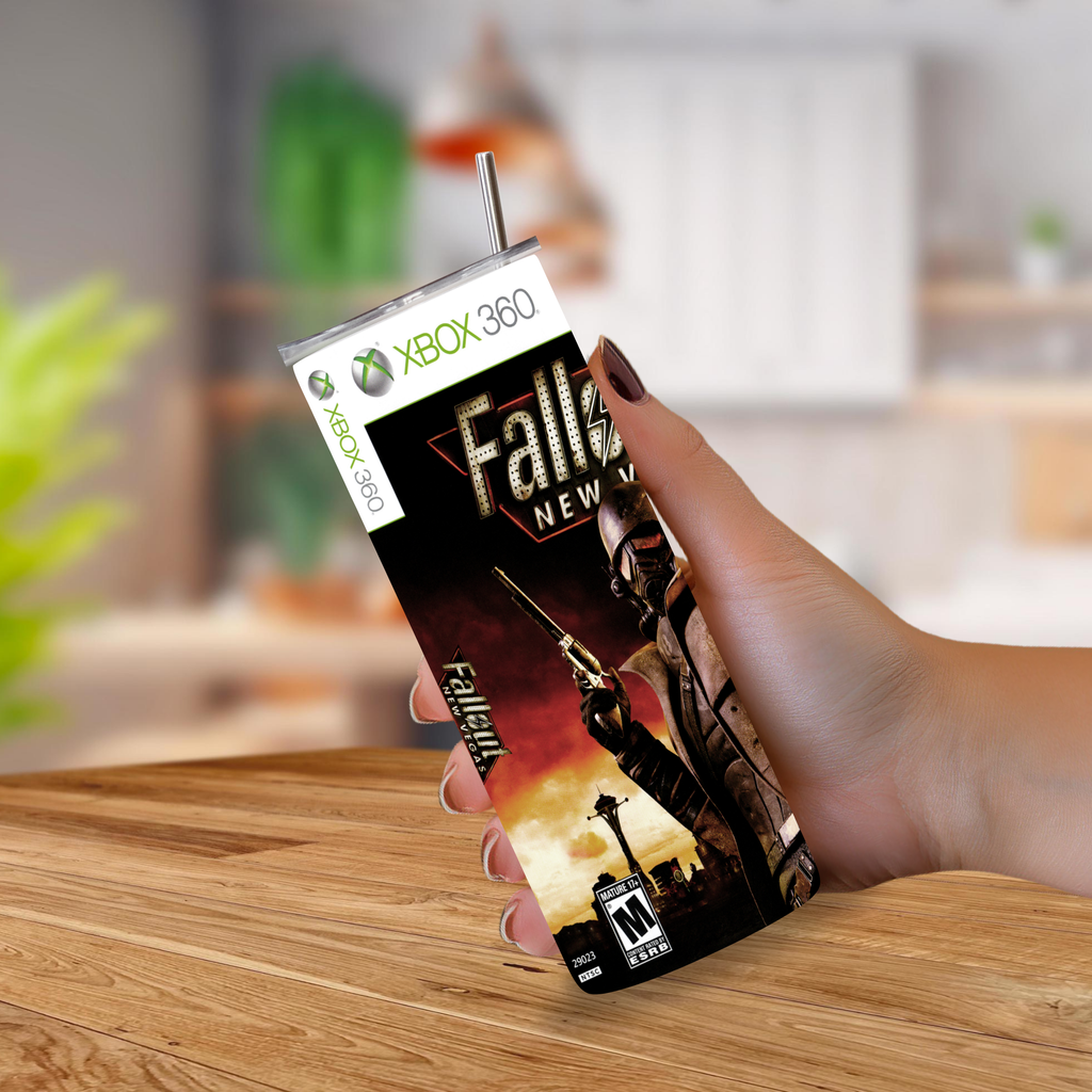 A hand holding a custom-printed tumbler cup designed to look like an Xbox 360 game case featuring the video game 'Fallout: New Vegas'. The cup has a clear plastic lid with a metal straw and is photographed on a wooden table with a blurred indoor background.